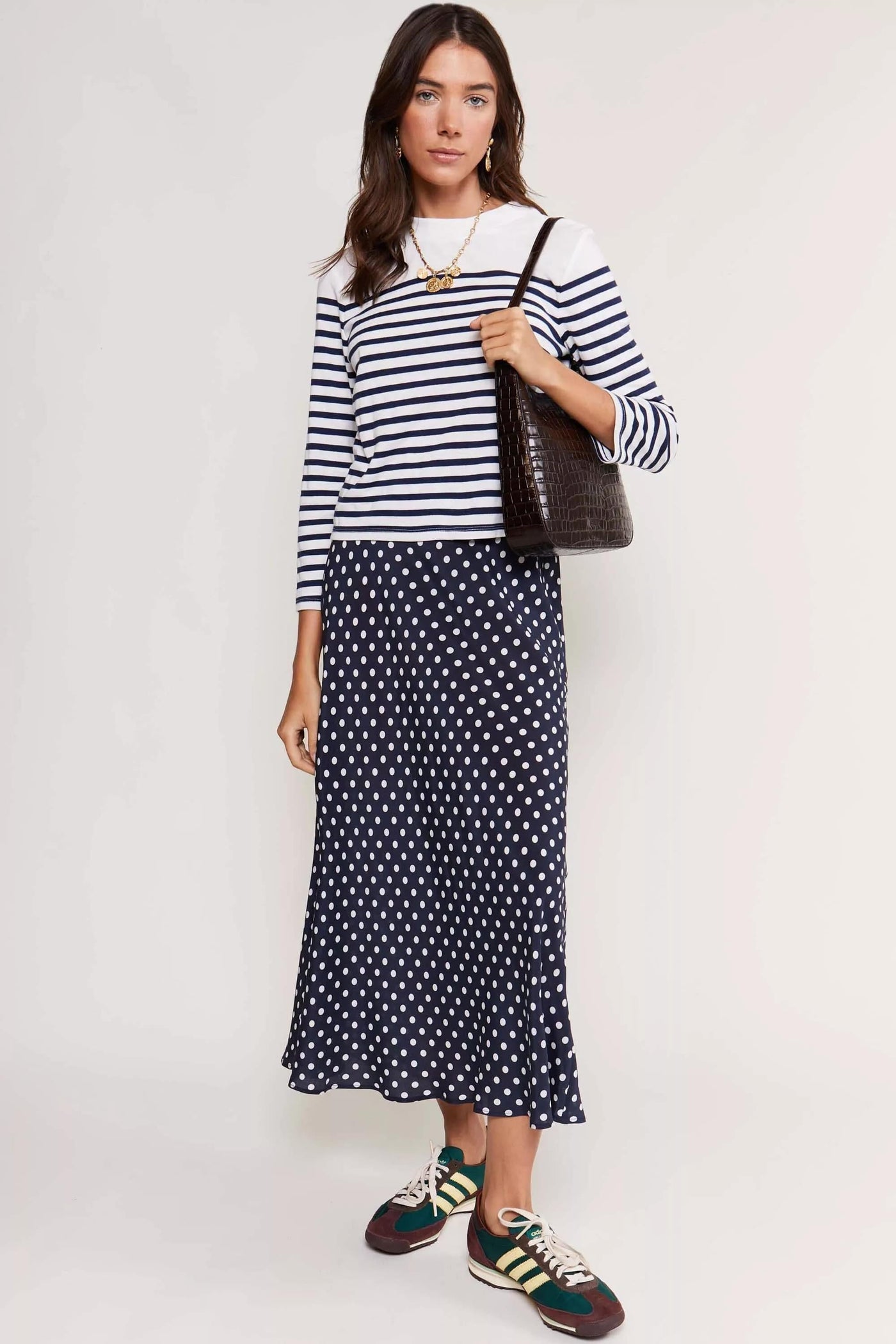 A woman stands against a white background wearing the Francesco - Nautical Stripe Cream top, a navy polka dot skirt, green and white sneakers, and carrying a dark brown textured shoulder bag—a timeless wardrobe staple look.
