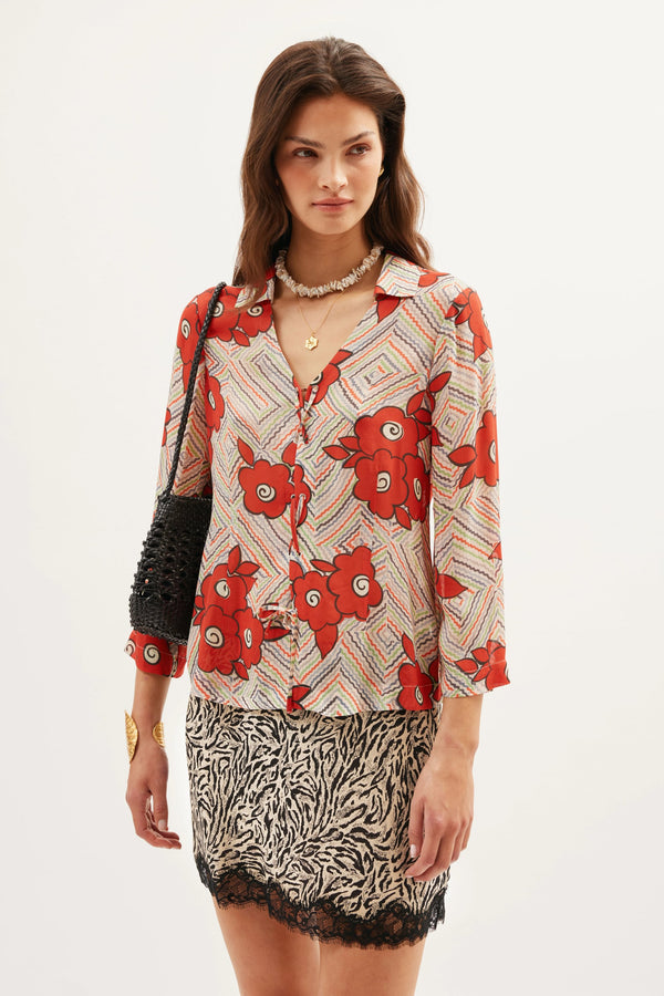 A woman stands against a plain background wearing a cream floral Henny Milos blouse, a black and white animal print skirt with lace trim, a black handbag, and layered necklaces.