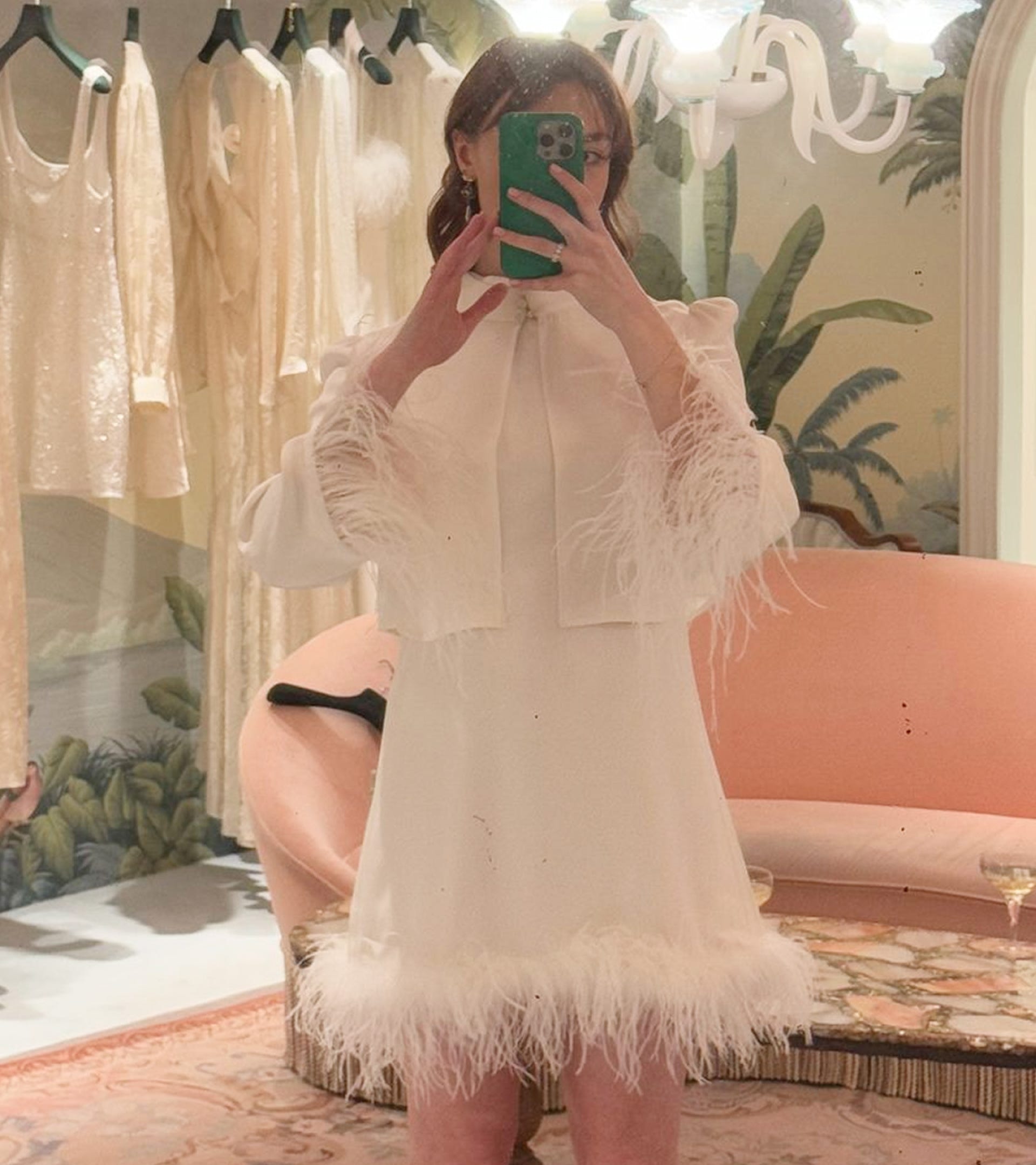 Person wearing a white dress with feather details in a room with floral wallpaper and a pink couch.