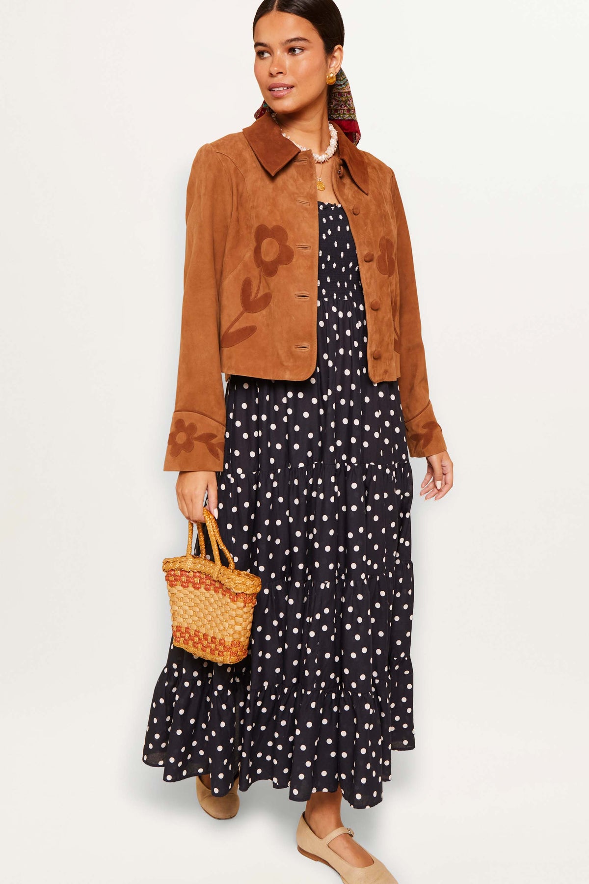 A woman models the Kendall - Bohemia Spot Deep Navy, a breezy polka-dot maxi dress, styled with a brown suede jacket, woven handbag, and tan shoes against a plain background.