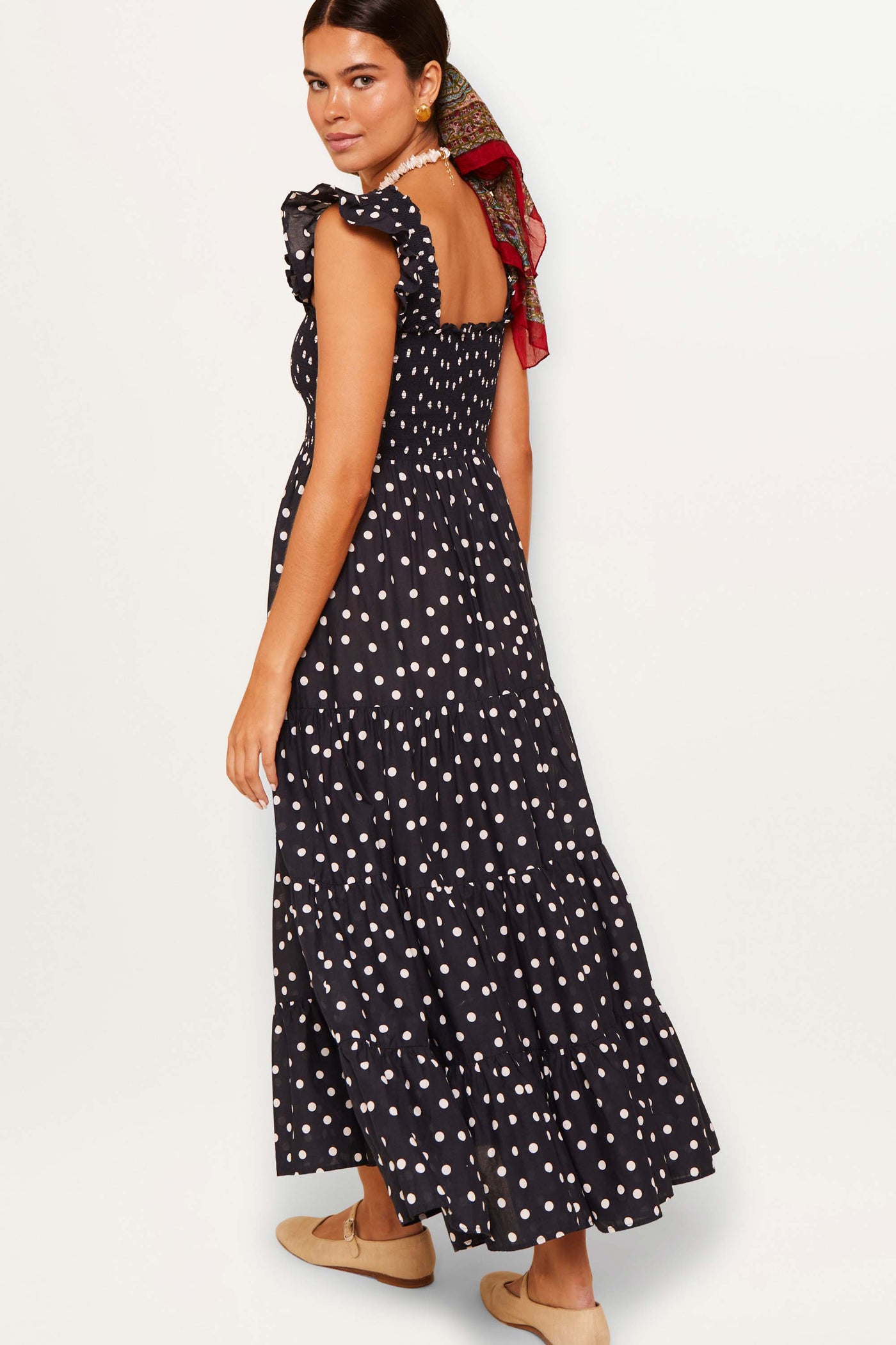 A woman in the Kendall - Bohemia Spot Deep Navy dress, featuring black cotton-voile with white polka dots and ruffled straps, stands turned slightly over her shoulder against a white background.