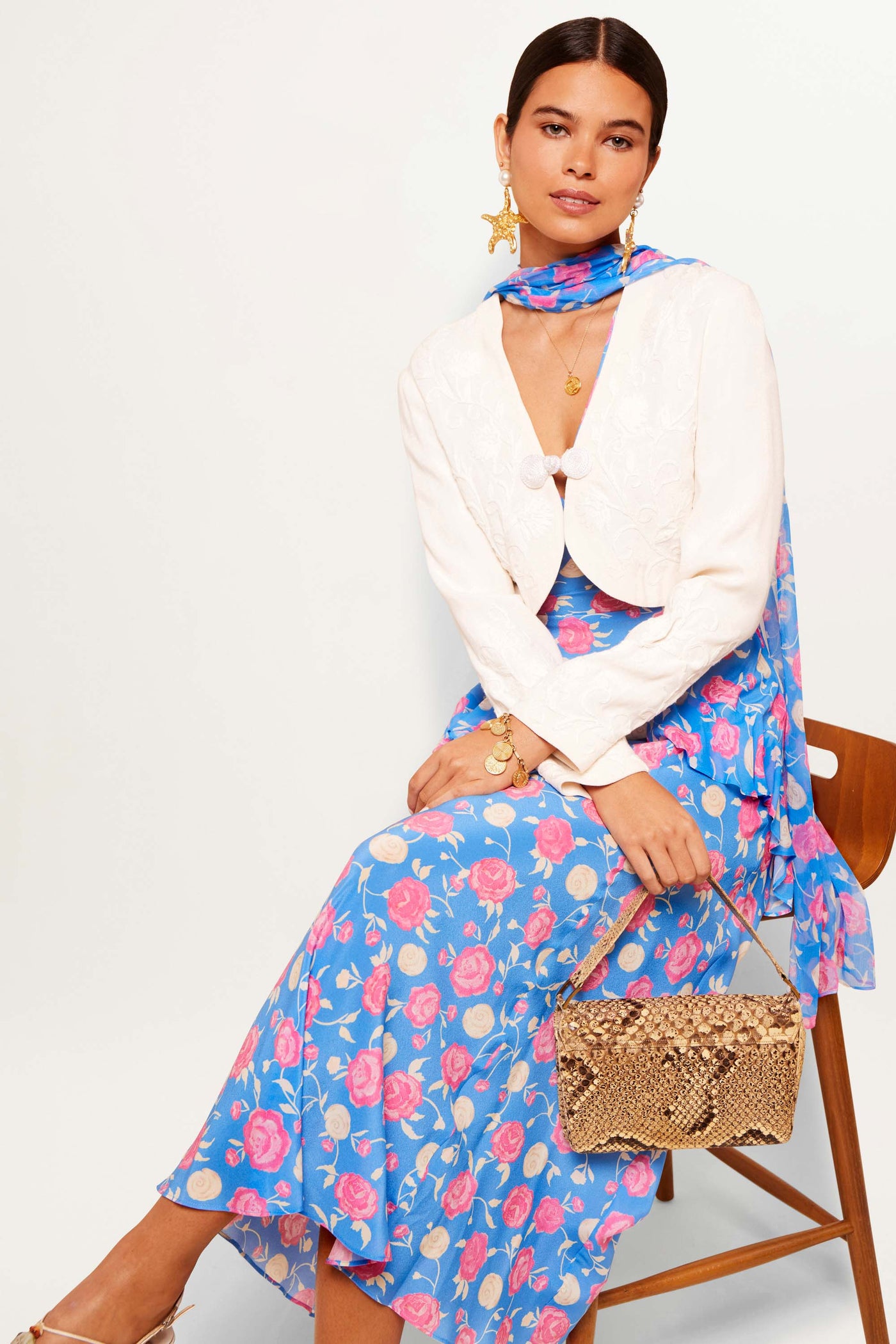 A woman sits on a wooden chair wearing the Evie - Rose Shell Blue dress, featuring white, pink, and orange floral patterns. She has on gold jewelry, carries a beige textured handbag, and her hair is pulled back.