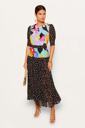 A woman stands against a white background wearing the Cozi - Tulip Spot Mix Multi: a colorful RIXO floral top and a black midi skirt with white polka dots, styled with a beige clutch, strappy heels, gold jewelry, and her hair pulled back.