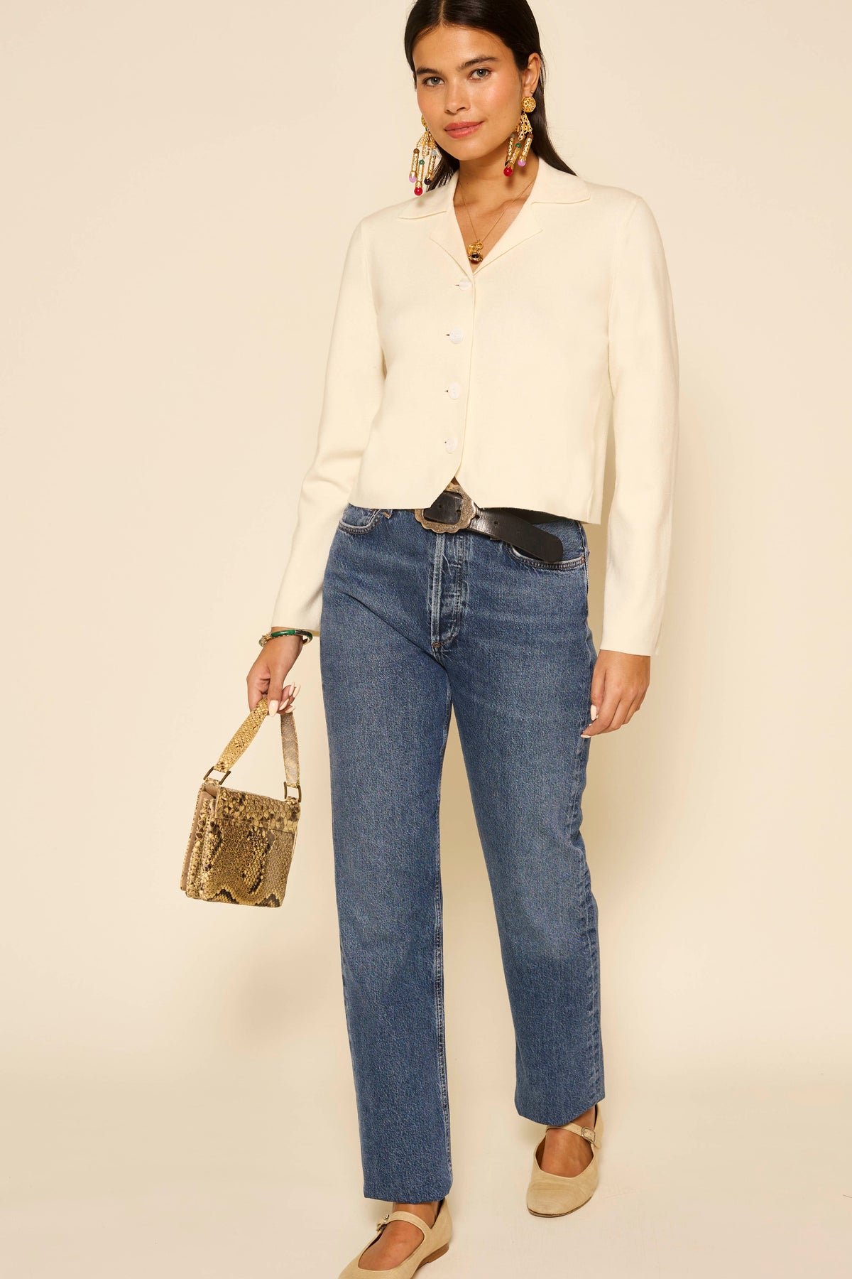 A woman with long dark hair and a confident expression stands against a beige background wearing the Aara - Ivory blouse, layered with a vintage waistcoat, styled with blue jeans, beige flats, statement earrings, and a small snakeskin-pattern handbag.