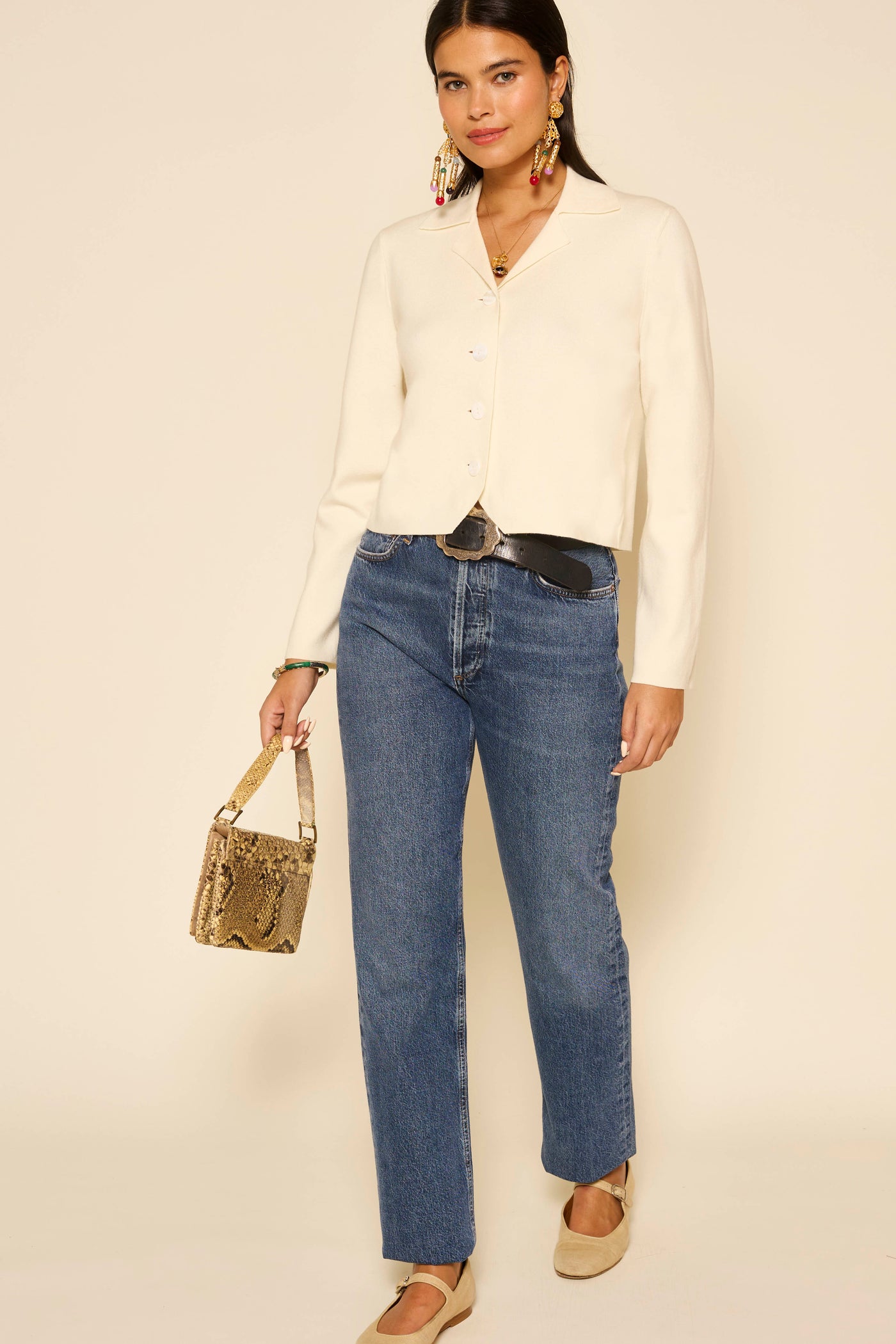 A woman with long dark hair and a confident expression stands against a beige background wearing the Aara - Ivory blouse, layered with a vintage waistcoat, styled with blue jeans, beige flats, statement earrings, and a small snakeskin-pattern handbag.
