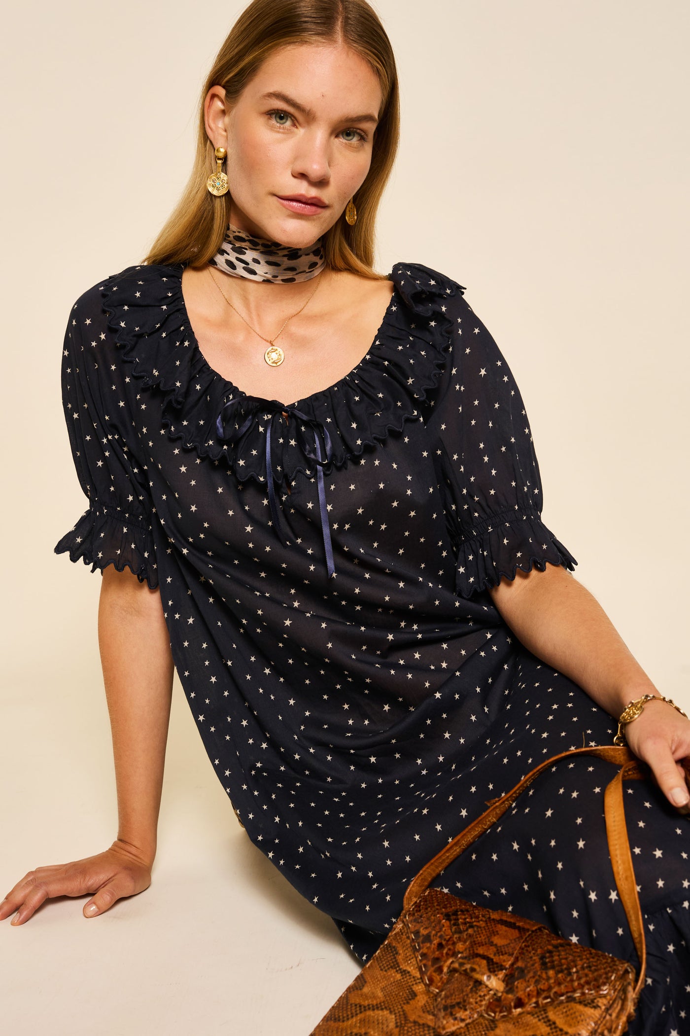 A woman with long blonde hair sits on the floor wearing the Aprilia - Starry Night Navy dress with a ruffled neckline, a patterned neck scarf, gold jewelry, and holding a brown bag.
