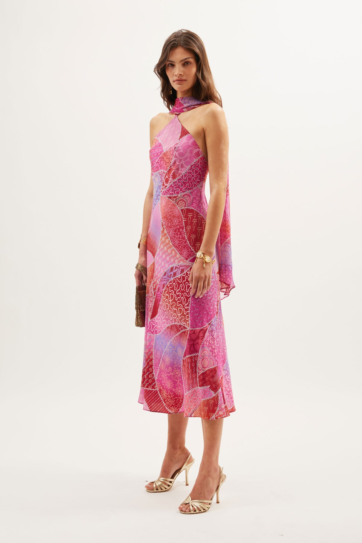 A woman stands against a plain white background in the Hayley - Ibiza Night Sunset dress, featuring a pink and purple silk crepe de chine halter-neck with a detachable georgette scarf, holding a clutch and wearing strappy heels.