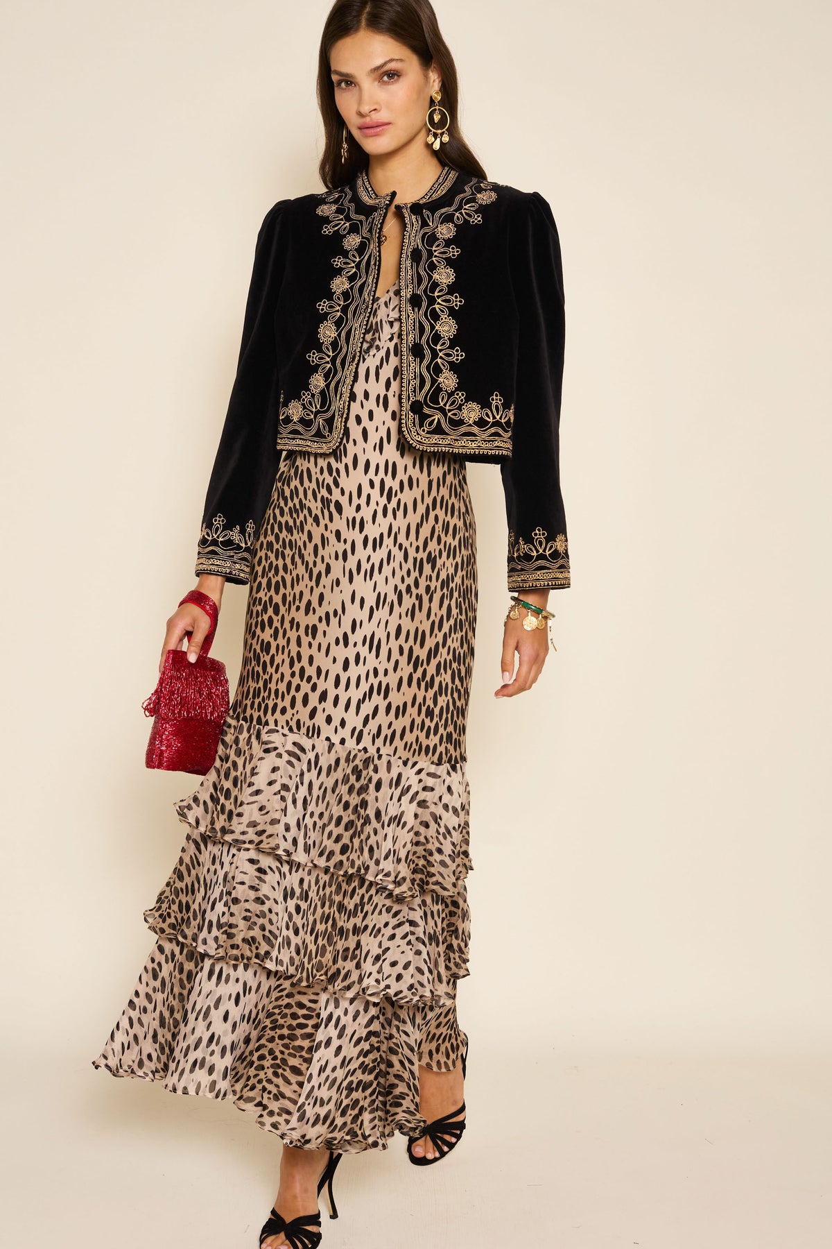 A woman stands against a plain background in the Amelle - Antelope Spot Black, a ruffled animal-print maxi dress with fluted sleeves. She pairs it with a black cropped jacket with gold embroidery, red textured handbag, statement earrings, and black heels.