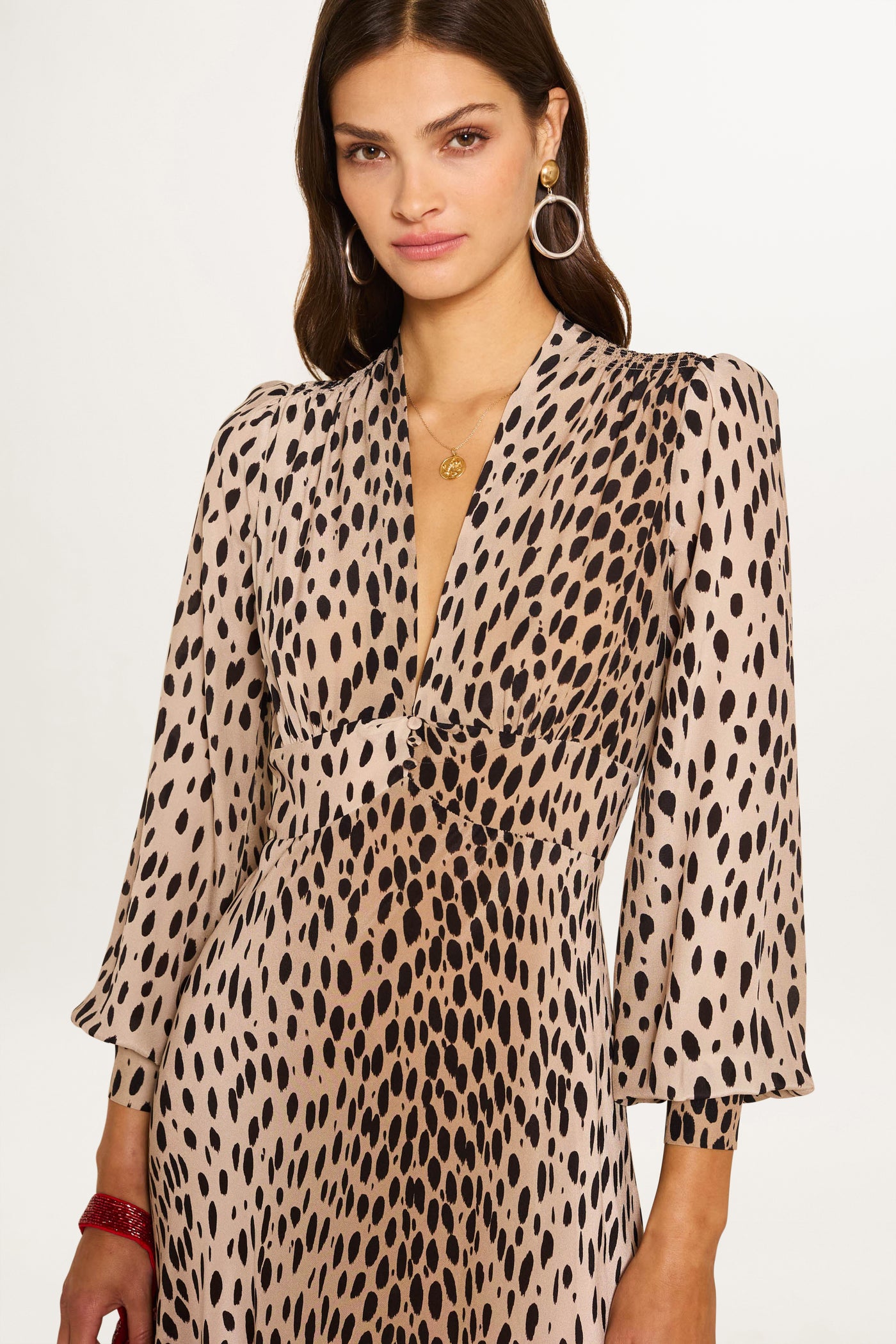 A woman with long brown hair wears the Emory - Antelope Spot Black dress, a beige silk crepe de chine with black spot print, long sleeves, and a deep V-neckline. She accessorizes with hoop earrings and a gold necklace against a plain white background.