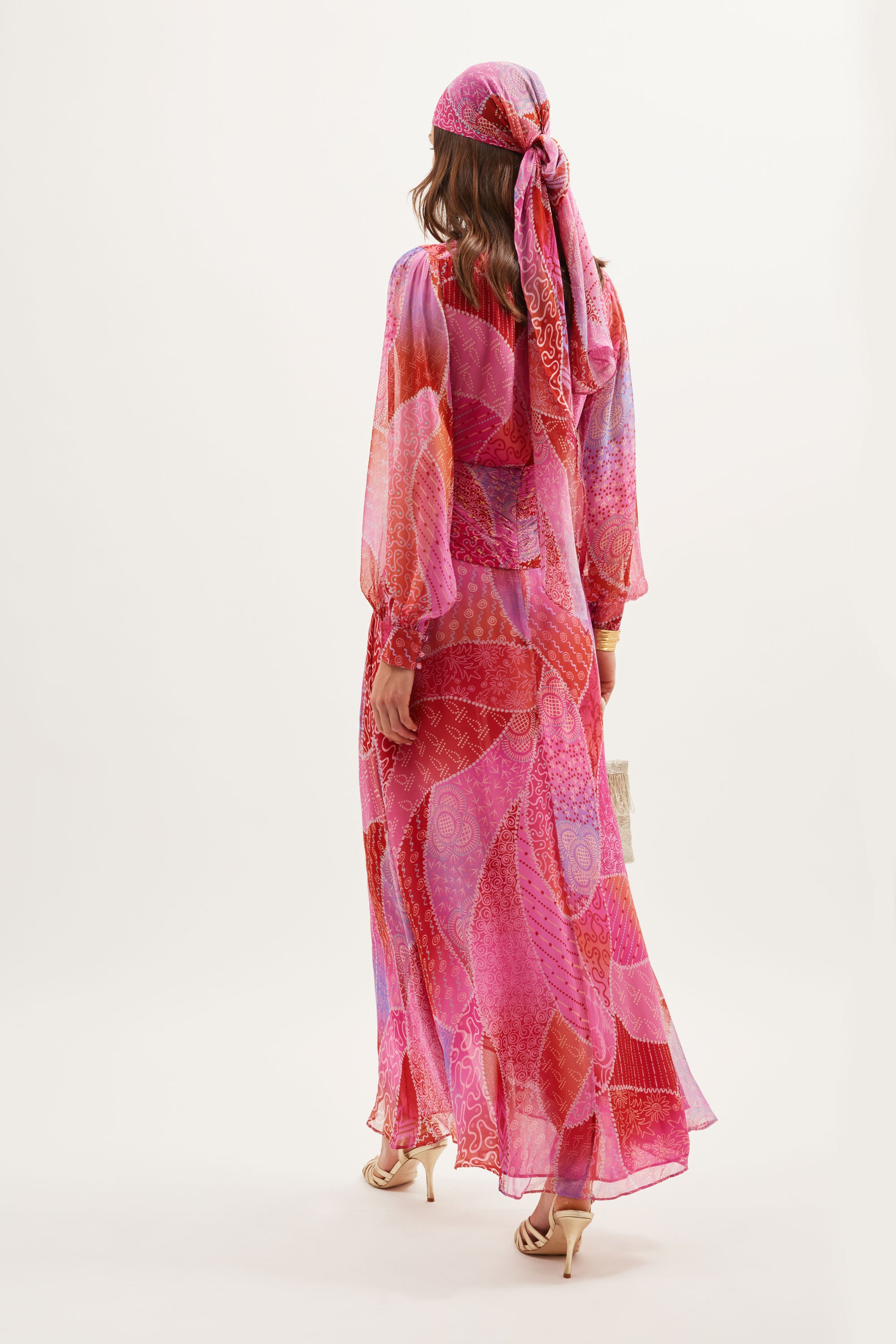 A woman faces away, dressed in the Meera - Ibiza Night Sunset gown, featuring crinkled-chiffon, long sleeves, and a matching headscarf. She carries a small bag and wears heeled sandals against a simple, light backdrop.