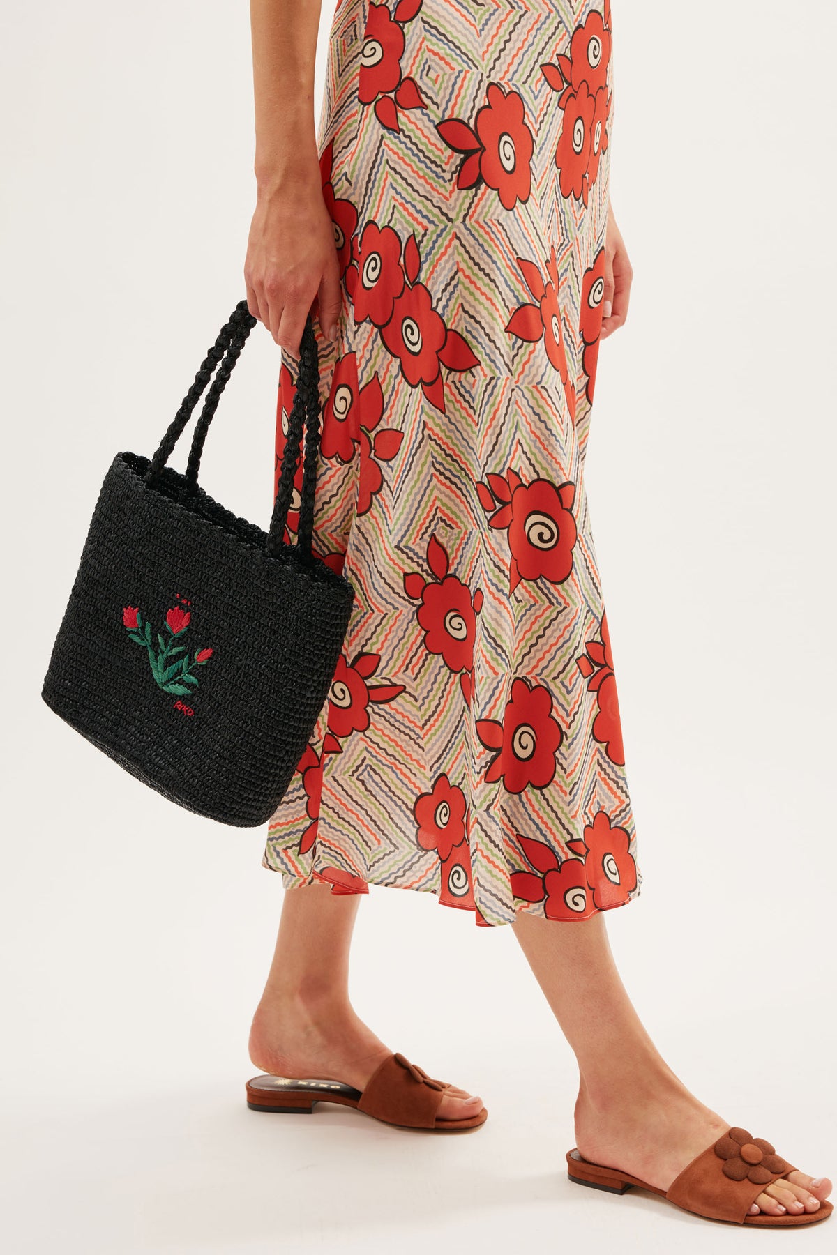 A person in a floral skirt and brown Kenna sandals holds a black woven handbag with red flower embroidery, capturing an effortless summer vibe. Only their lower half is shown.