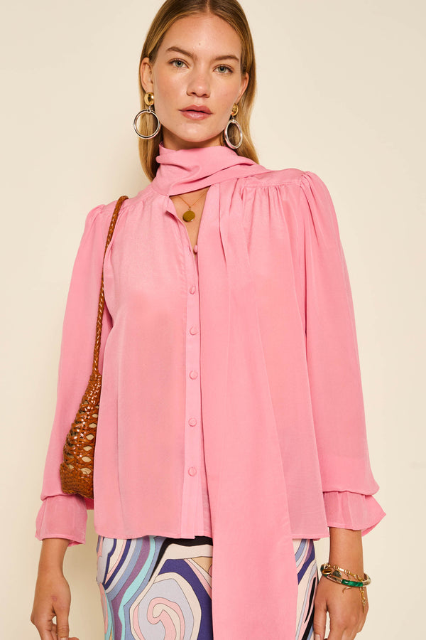 A woman wearing the Wilhelmina - Blush Pink pussy-bow blouse, patterned pastel pants, gold hoop earrings, and a brown woven bag stands against a plain background.