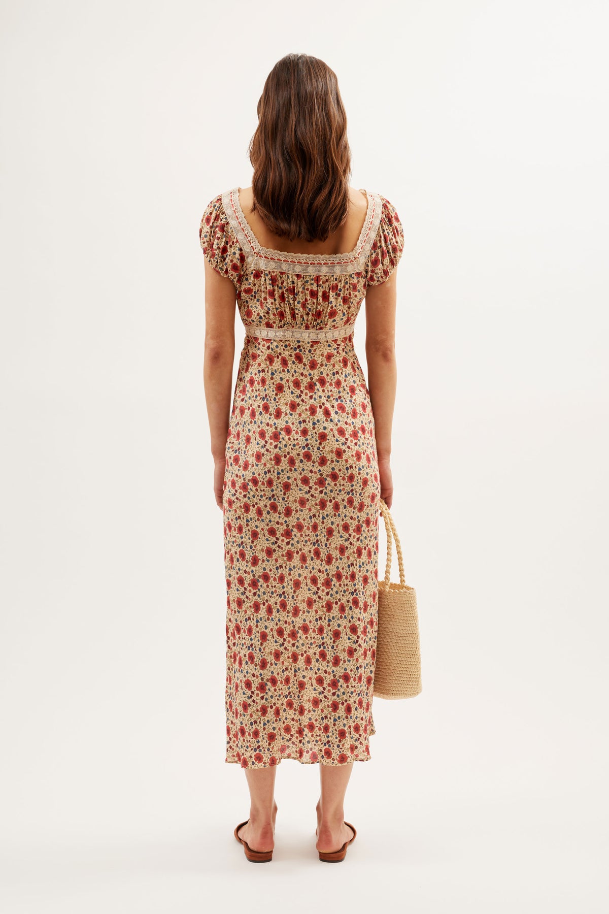 A woman with brown hair, seen from behind, wears the Maeva - Eva Floral Cream ankle-length dress with a square neckline and lace trim. She holds a straw handbag in her left hand and stands against a plain white background.