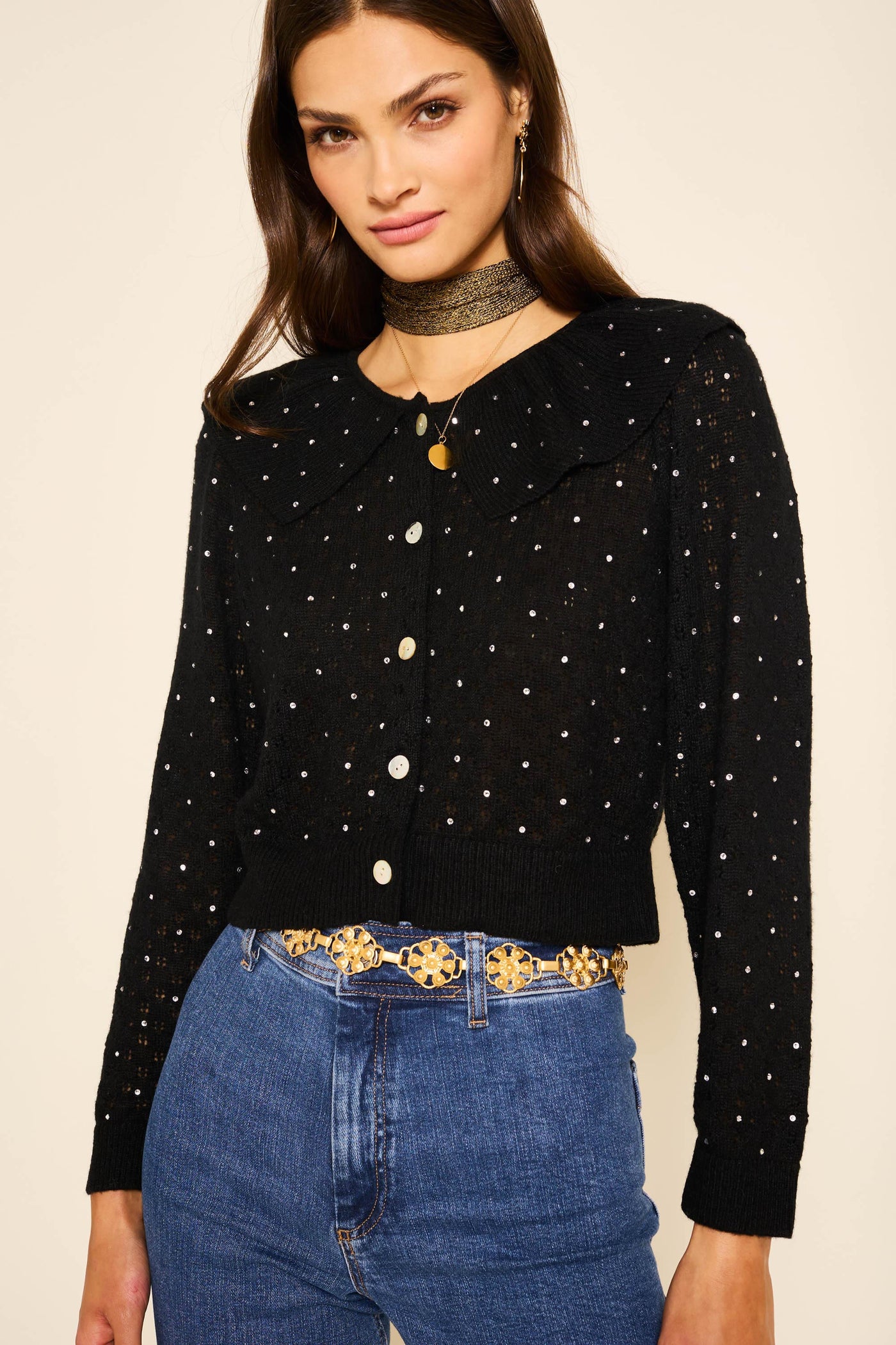 A woman wears the Jelena - Crystal Black long-sleeve, pointelle-knit button-up sweater with white dots, paired with blue jeans, a black choker, and a gold belt, standing against a light background.