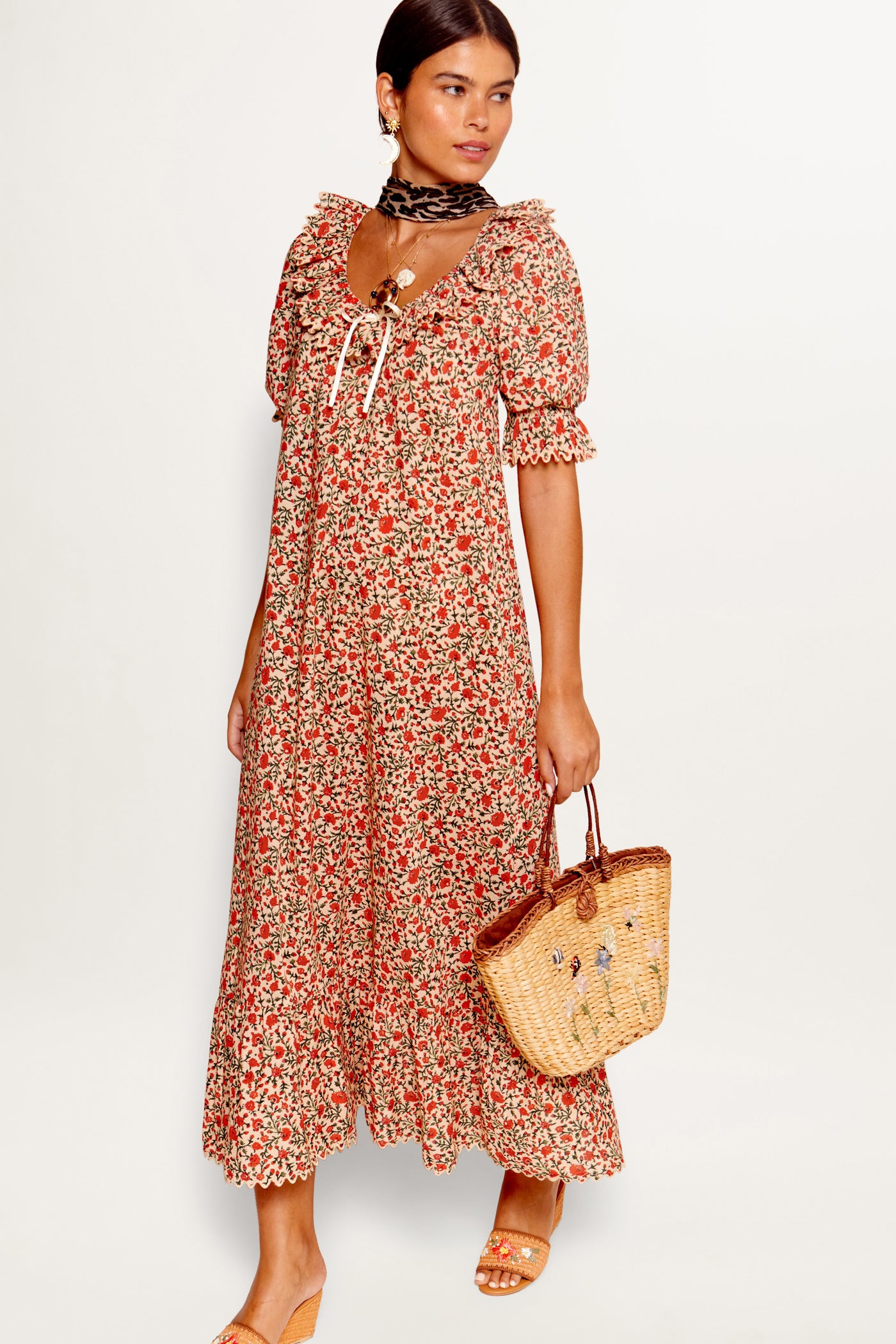 A woman wears the Aprilia - Ditsy Trail Cream long dress with ruffled sleeves, paired with sandals, a woven straw bag, hoop earrings, and a patterned neck scarf against a white background.