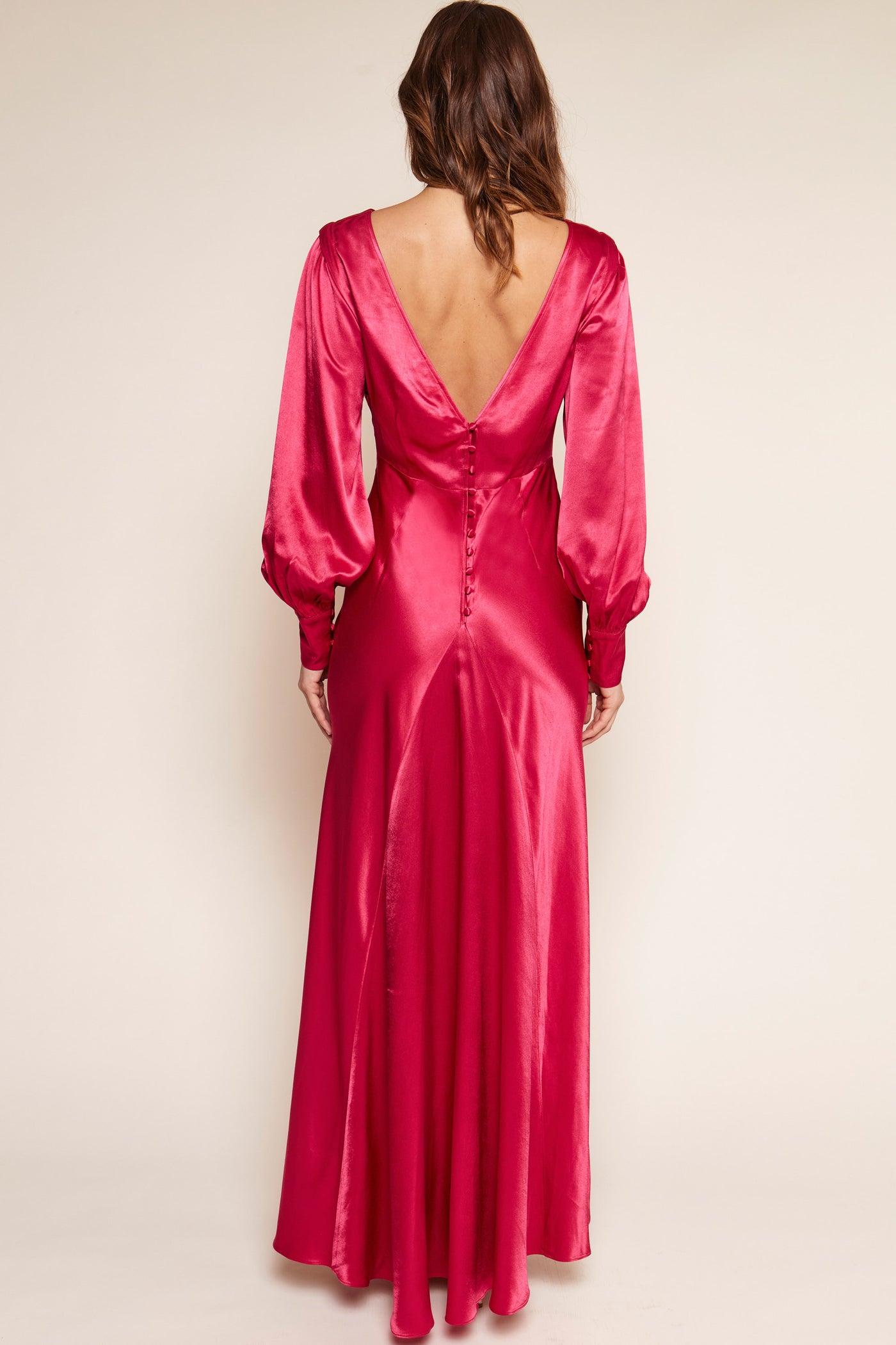 A woman with wavy brown hair wears the Marni - Raspberry, a floor-length vintage-inspired gown in vibrant hot pink satin, featuring a deep V-back and button details, photographed from behind against a plain light background.
