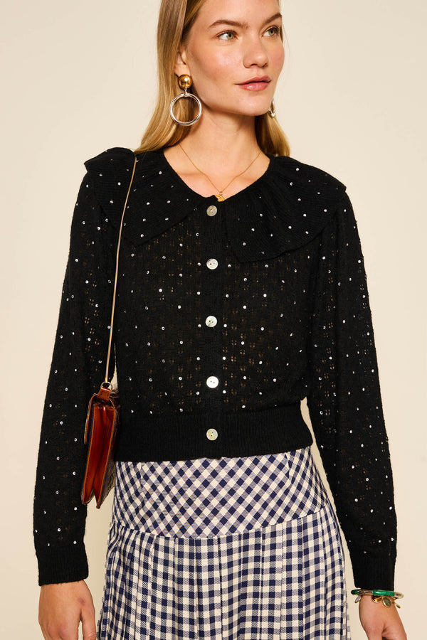 A woman wears the Jelena - Crystal Black party cardigan with white polka dots, a large collar, and pairs it with a blue and white checkered skirt. She carries a brown shoulder bag and accessorizes with hoop earrings.