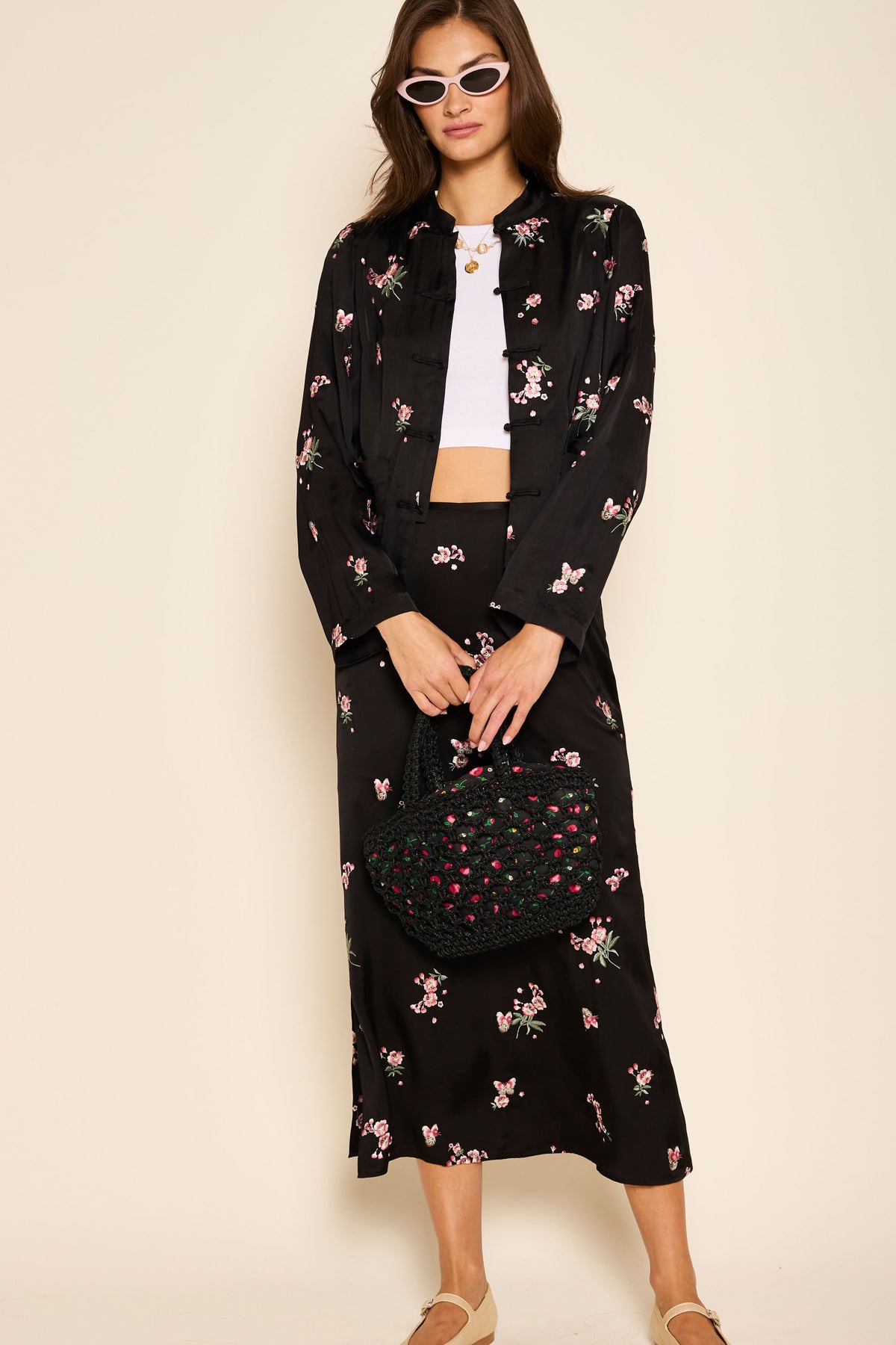 A woman wears the Indiya - Blossom Embroidery Pink skirt in a column silhouette with a matching jacket, white crop top, sunglasses, beige shoes, and holds a black woven handbag with pink accents. Regular and petite sizes available.