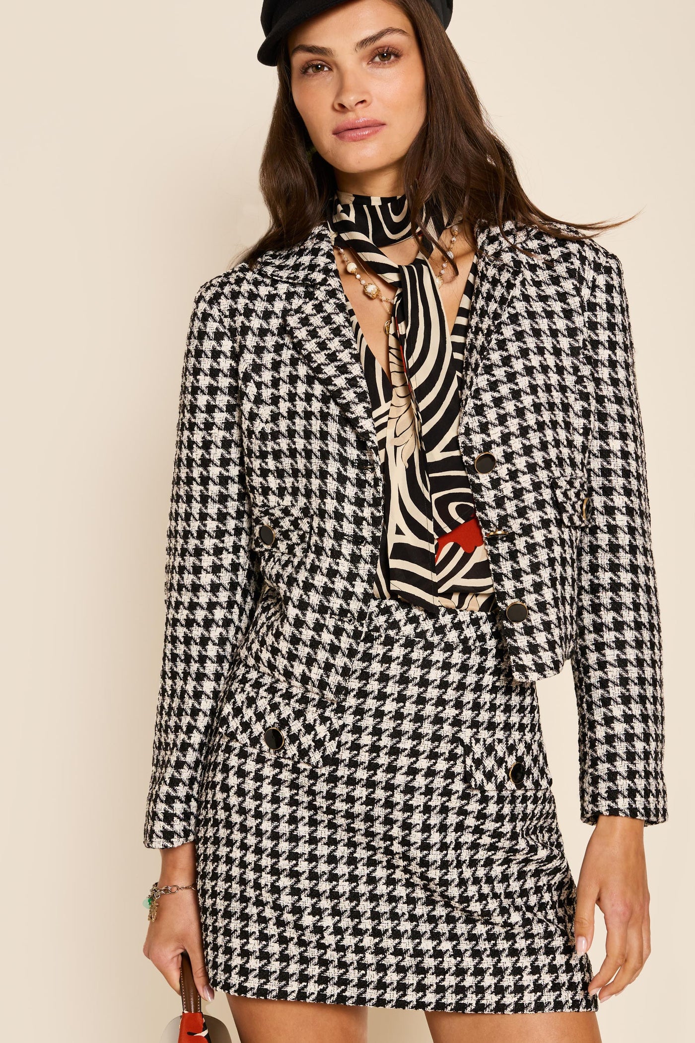 A woman wears the Mariah - Houndstooth Mono mini skirt and matching blazer, styled with a noughties blouse, scarf, black beret, and small bag, standing against a beige background.