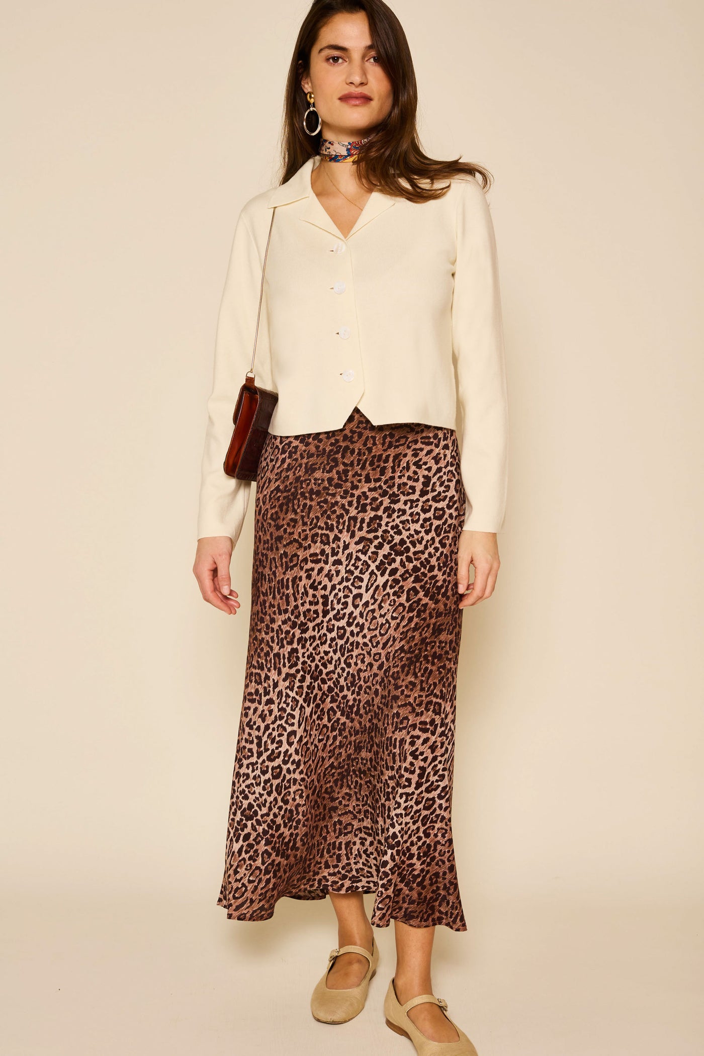 A woman stands against a beige background, wearing the Aara - Ivory cream merino wool-blend cardigan, a brown leopard print midi skirt, beige Mary Jane shoes, hoop earrings, a choker scarf, and carrying a small brown shoulder bag.