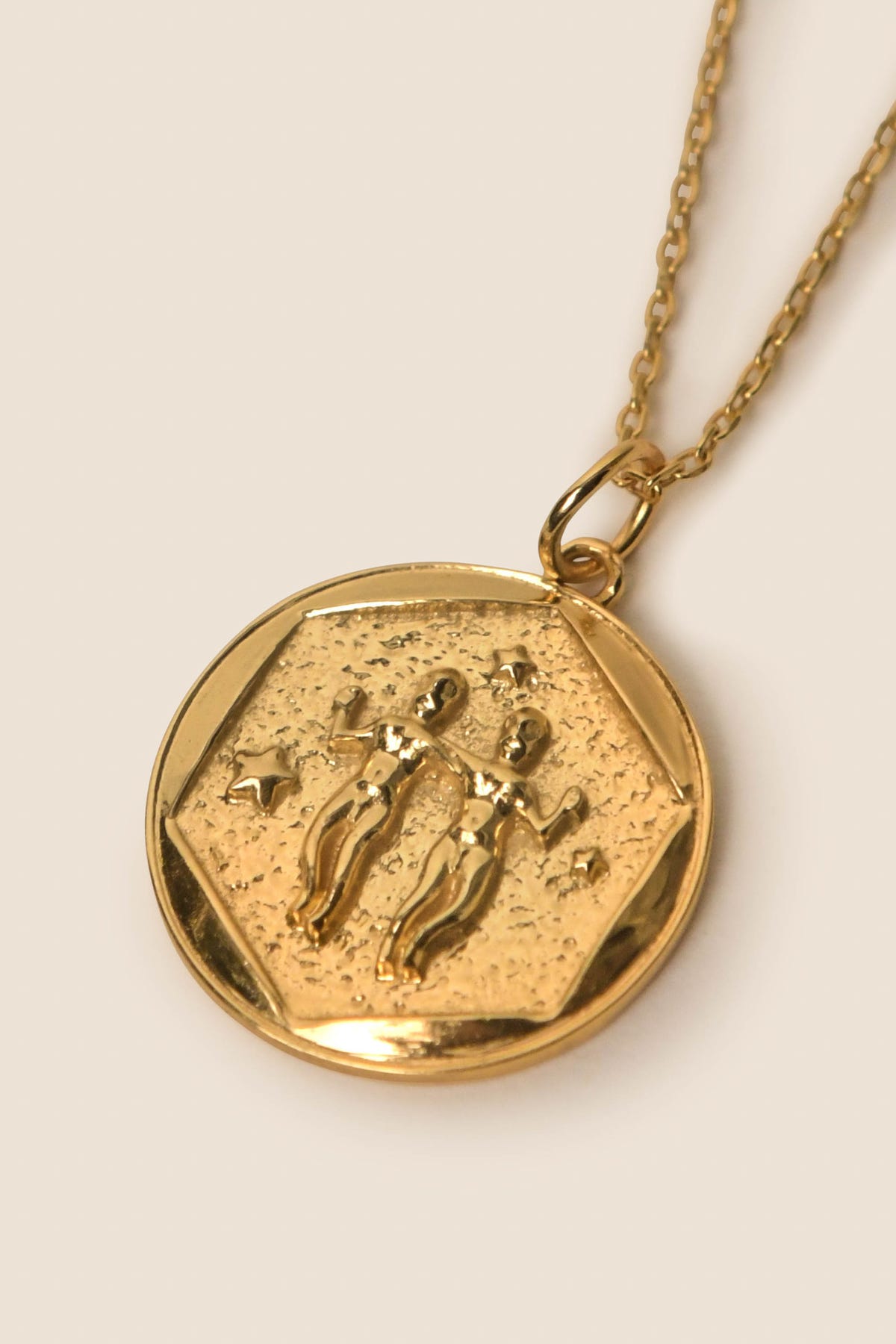 The Zodiac - Gemini necklace features a gold vermeil round pendant with two embossed human figures symbolizing Gemini, surrounded by stars on a textured background—a perfect accessory for Zodiac jewelry enthusiasts.