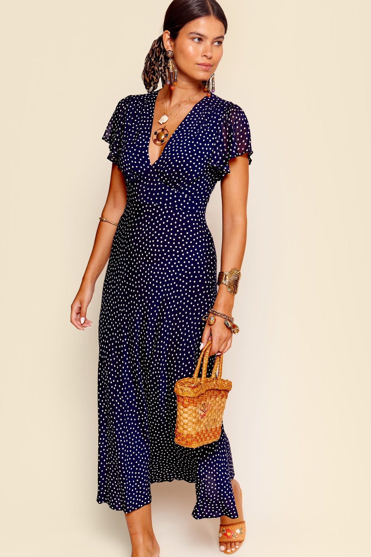 A woman wears the Florida - Mini Dot Navy dress, a navy blue, short-sleeved fit and flare style with polka dots. She holds a woven straw handbag and accessorizes with statement earrings, bangles, sandals, and a low ponytail.