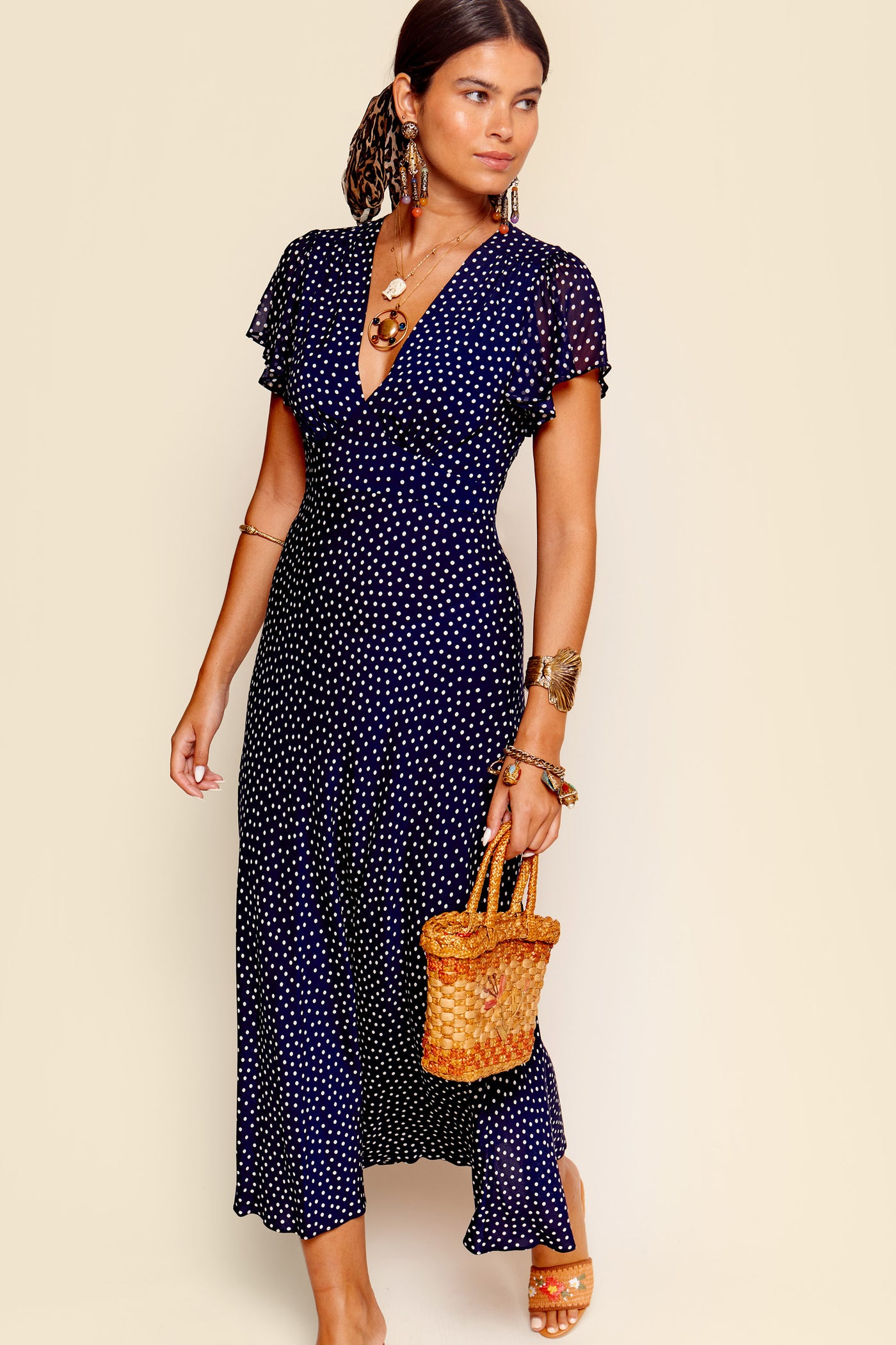 A woman wears the Florida - Mini Dot Navy dress, a navy blue, short-sleeved fit and flare style with polka dots. She holds a woven straw handbag and accessorizes with statement earrings, bangles, sandals, and a low ponytail.