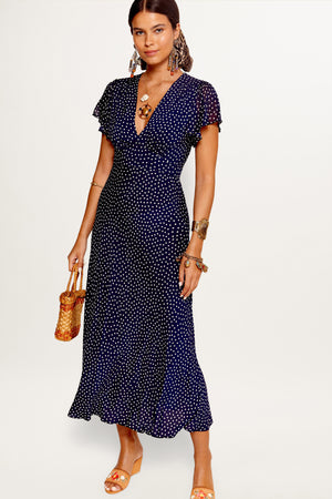 A model wearing a navy dress with a mini polka dot pattern, featuring a V-neck, short sleeves, and a midi length.