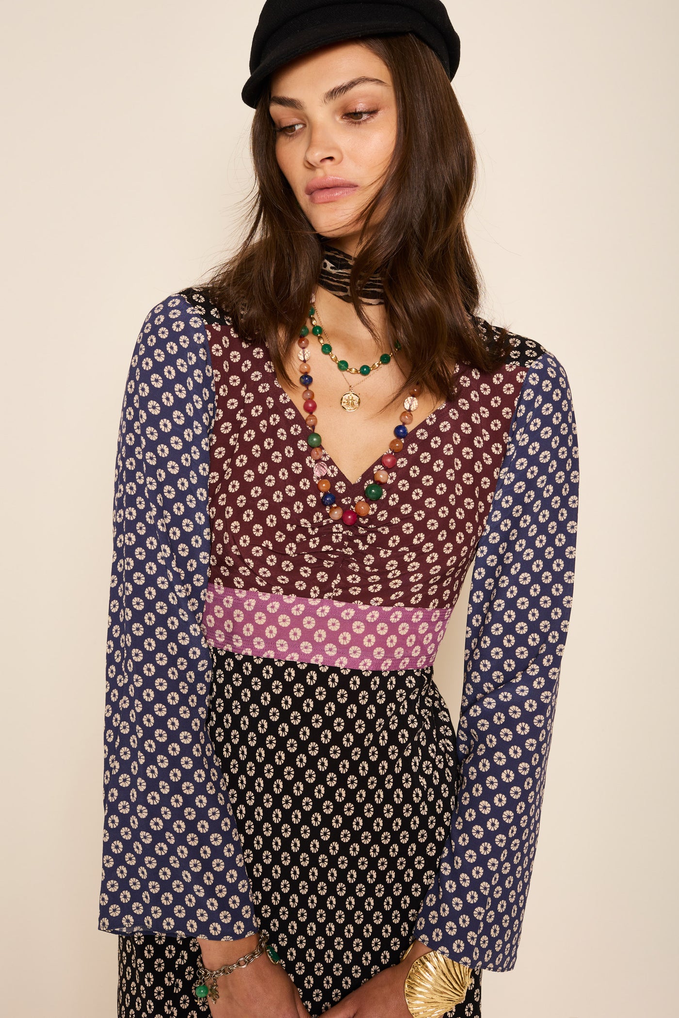 A woman wears the Priscilla - Shibori Burgundy Mix dress—featuring blue sleeves, a maroon bodice, and black-and-white skirt with detachable waist ties—styled with layered necklaces, bracelets, and a black beret against a plain background.