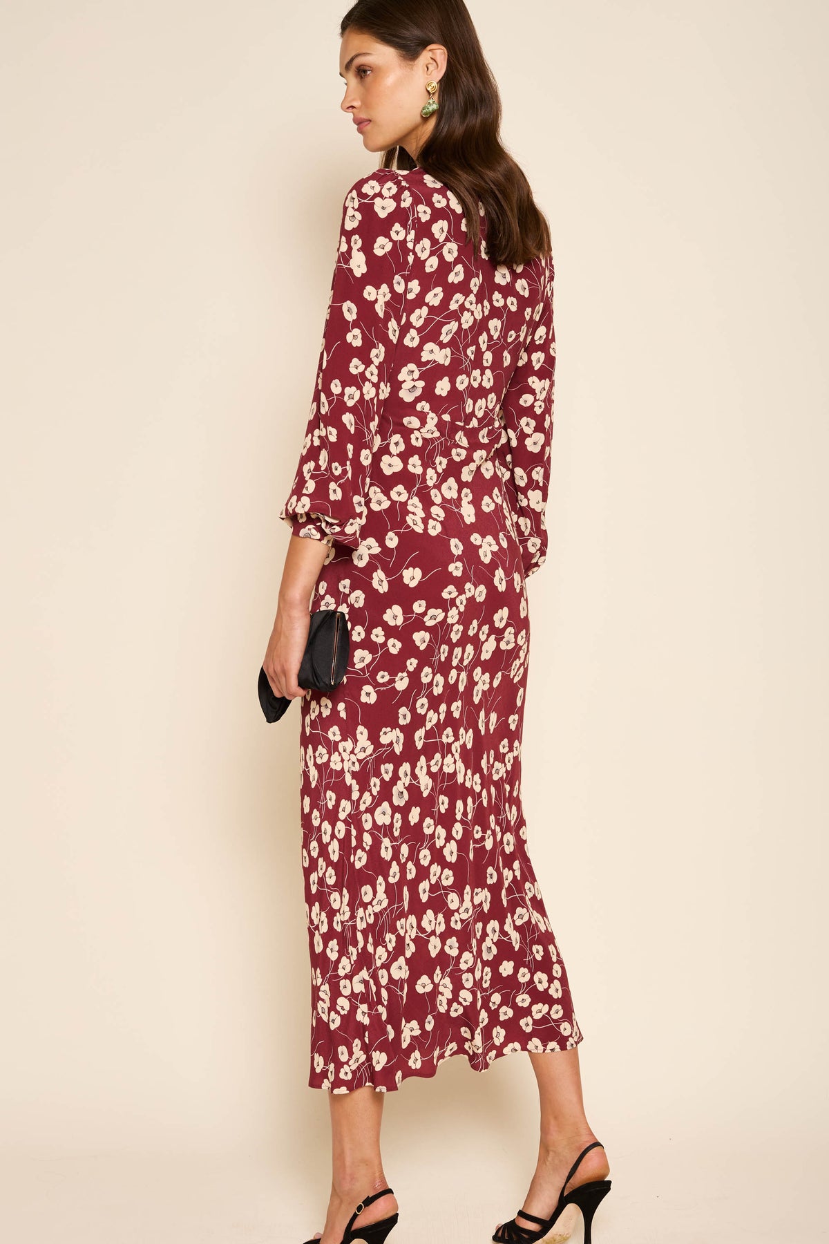 A woman stands facing away, wearing the Mavis - Autumn Cherry Maroon dress with everyday florals. She holds a black clutch, wears black high-heeled sandals and gold earrings, and her brown hair is styled in loose waves.
