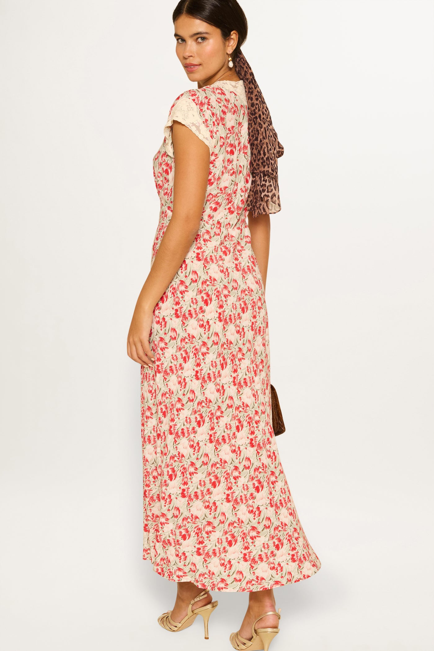 A woman in the Amari - Tulip Fields Red vintage-inspired maxi dress stands sideways, looking over her shoulder. She pairs it with beige heeled sandals and a leopard-print hair scarf against a plain white background.