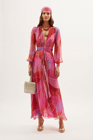 A woman wears the Meera - Ibiza Night Sunset, a colorful long crinkled-chiffon gown with pink, red, and purple patterns, long sleeves, a matching headscarf, gold sandals, layered necklaces, and a beige handbag against a white background.
