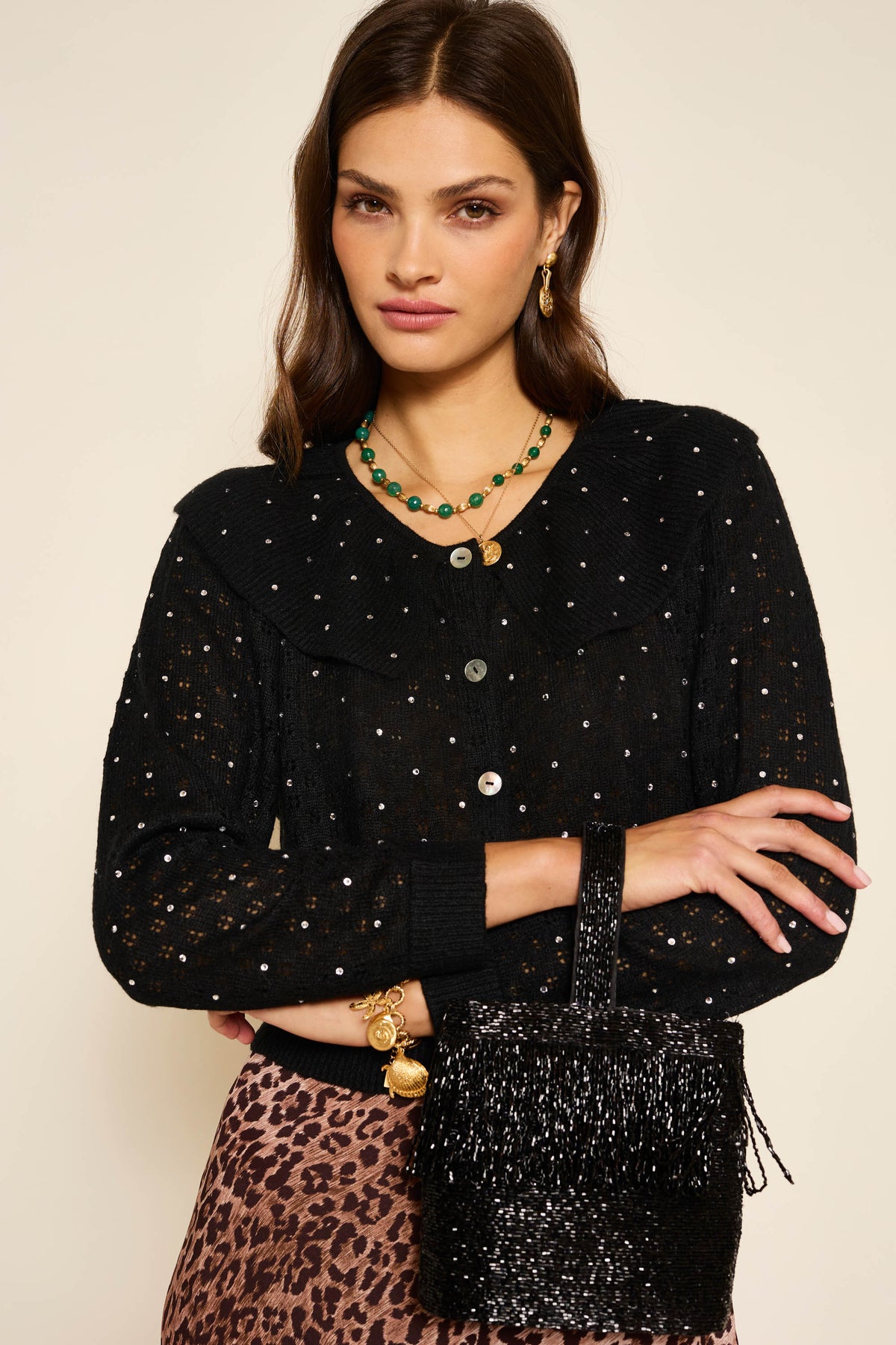 A woman with long brown hair wears the Jelena - Crystal Black pointelle-knit cardigan with small white dots, paired with a green beaded necklace, gold jewelry, a black beaded fringe bag, and a leopard-print skirt.