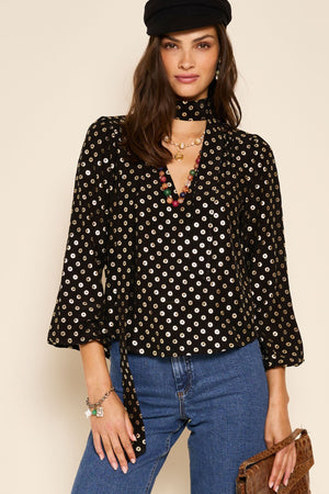 A woman with long dark hair and a confident expression wears Moss - Shibori Gold, pairing it with a black hat, blue jeans, layered necklaces, a brown clutch, and a bracelet.