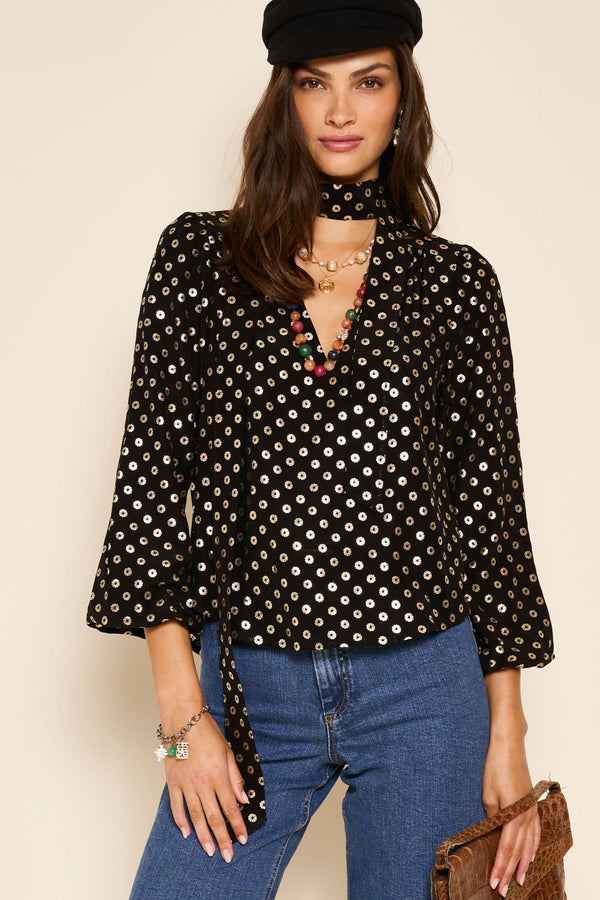 A woman with long dark hair and a confident expression wears Moss - Shibori Gold, pairing it with a black hat, blue jeans, layered necklaces, a brown clutch, and a bracelet.
