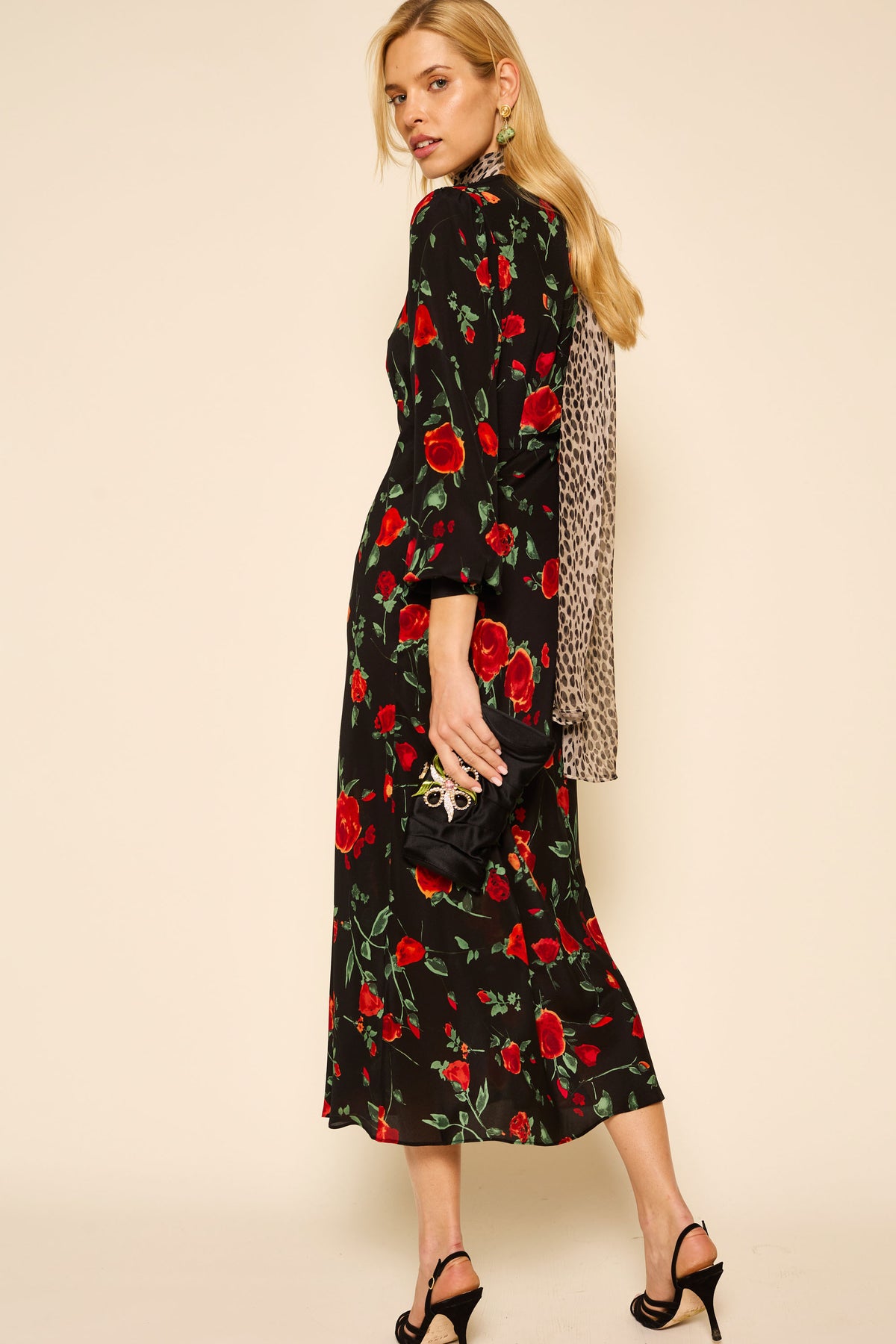 A woman with long blonde hair poses in the Mavis - Claudia Rose Black dress with a red floral pattern, black heeled sandals, a black clutch, and a patterned scarf draped down her back. She stands sideways, looking over her shoulder.