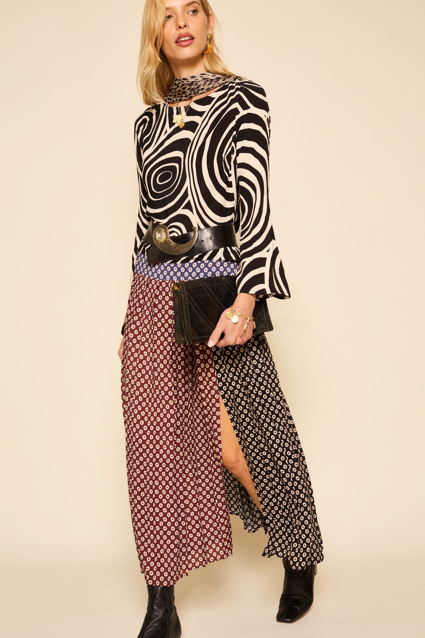 A woman stands against a beige background wearing the Verona - Whirlpool Mono top with fluted sleeves and a black-and-white swirl pattern, paired with a black wide belt, a long red and black geometric skirt, black ankle boots, and a black clutch.