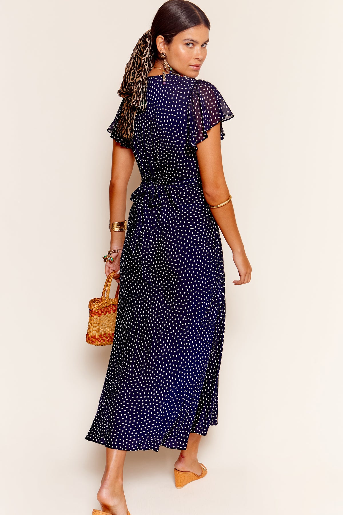 Wearing the Florida - Mini Dot Navy dress, a woman in tan sandals carries a woven handbag and wears gold jewelry. With her ponytail tied with a scarf, she looks over her shoulder—ideal for occasion wear or as a chic bridesmaid dress.