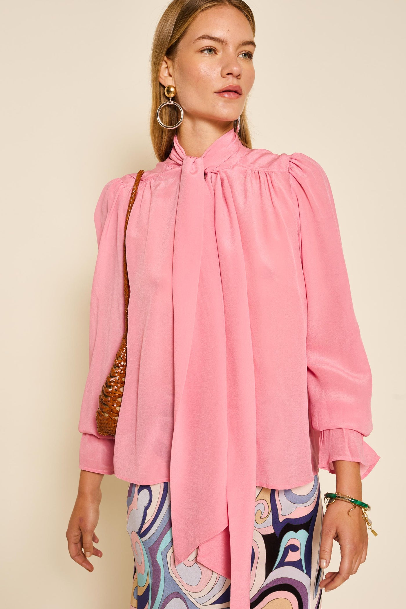 A woman wears the Wilhelmina - Blush Pink vintage-inspired pussy-bow blouse with puffed sleeves, a colorful abstract skirt, large hoop earrings, and a brown woven shoulder bag.