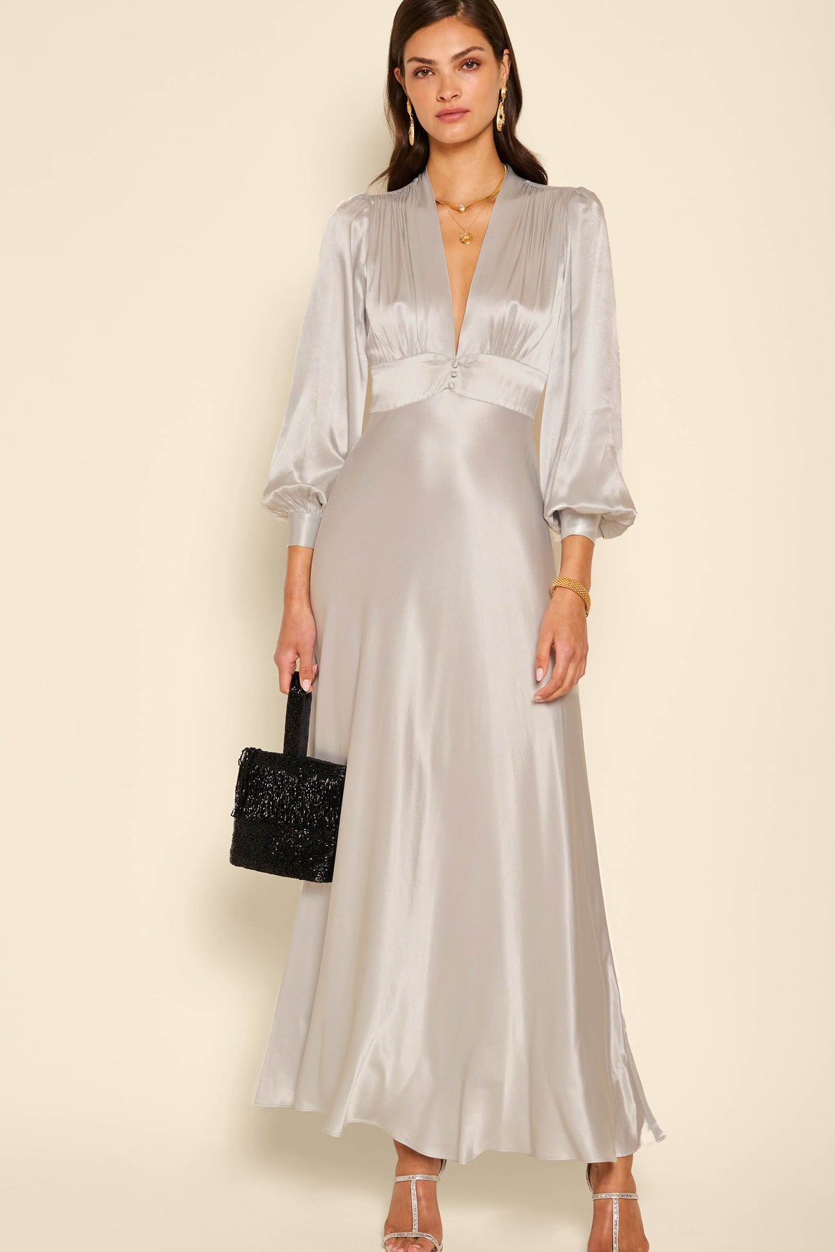 A woman wearing the emory - silver satin dress with voluminous shoulders, a deep V-neckline, and three-quarter sleeves holds a black textured handbag against a plain beige background.
