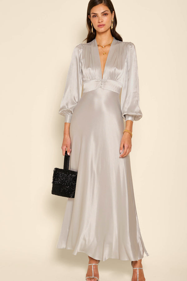 A woman wearing the emory - silver satin dress with voluminous shoulders, a deep V-neckline, and three-quarter sleeves holds a black textured handbag against a plain beige background.