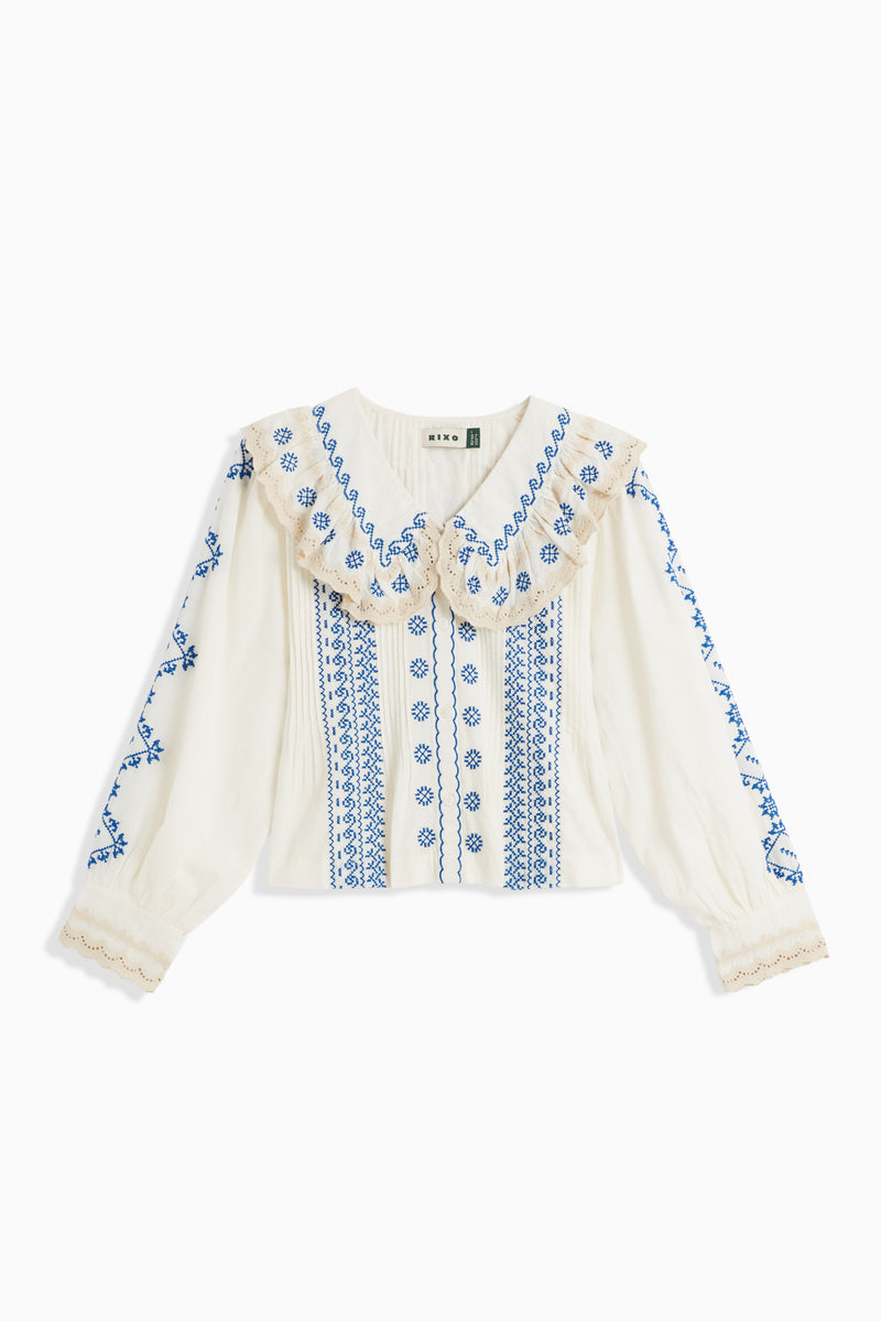 The Percy - Mila Embroidery Cream blouse features blue floral embroidery, a wide ruffled collar, beige trim, and intricate patterns down the front and sleeves for an effortlessly timeless vintage look.