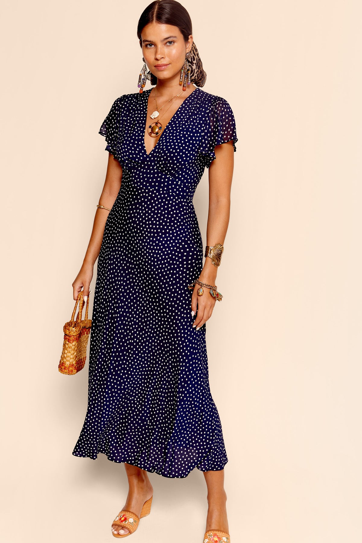 A woman wears the Florida - Mini Dot Navy dress, a short-sleeved, fit and flare polka dot midi. She pairs it with a woven handbag, statement jewelry, and tan wedge sandals while standing against a beige background.