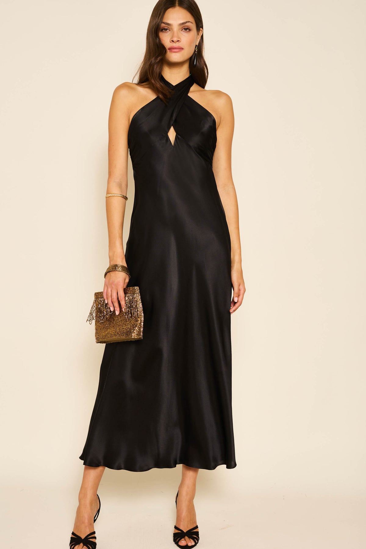 A woman stands against a light background wearing the Dune - Black sleeveless halter-neck silk-satin gown with a keyhole cutout, styled with a gold clutch, black heels, gold bangles, and hoop earrings.