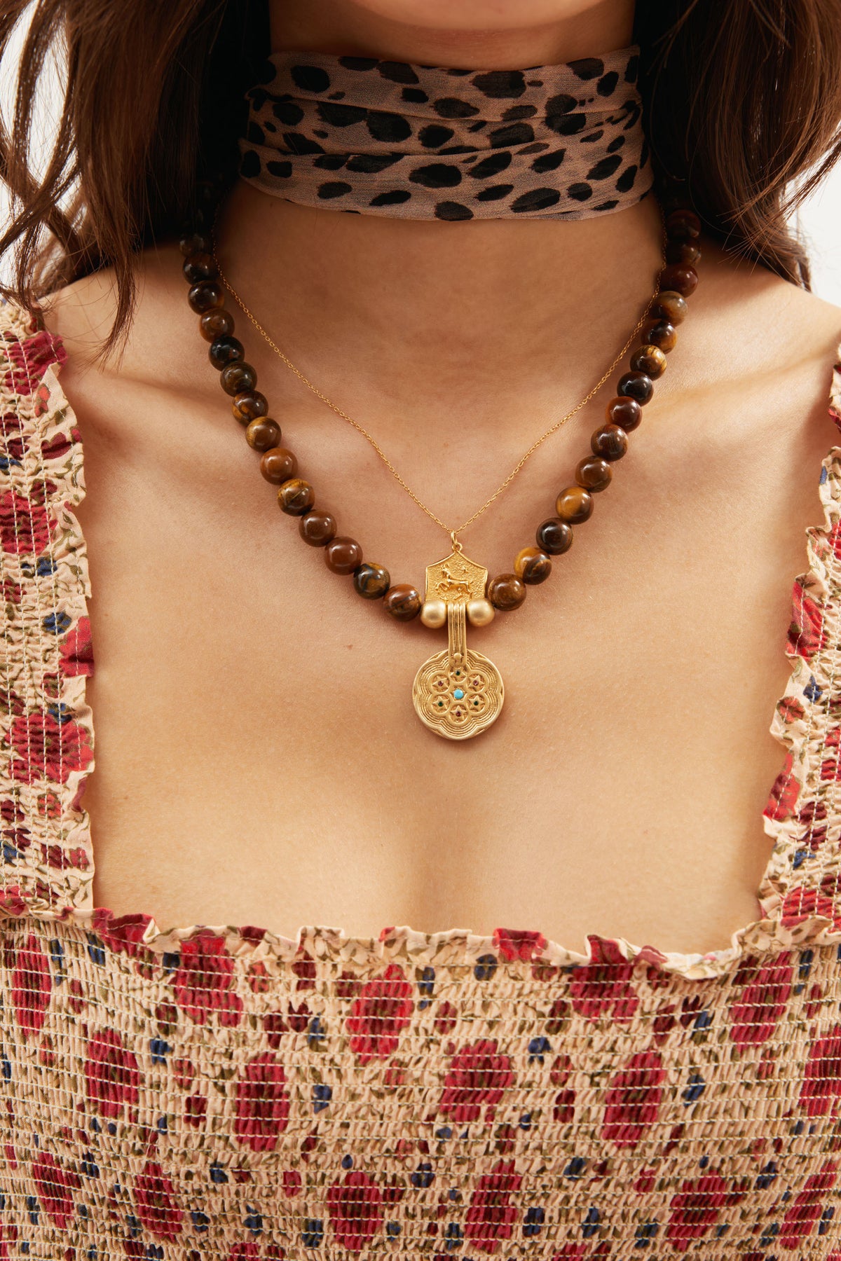 A woman wears the Ischia - Brown, a leopard-print choker, a beaded necklace with tiger's eye beads and gold pendant, and a gold chain. Her red and beige floral ruched dress is visible at the neck and chest.