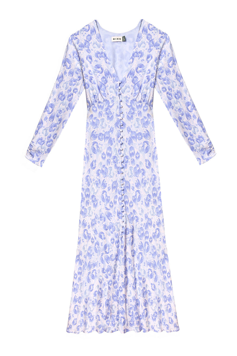 The Katie - Tulip Fields Cornflower Blue dress is a long-sleeve, V-neck, button-down midi with a fitted bodice and flared skirt, featuring light blue and lavender florals on white—ideal for spring and summer.