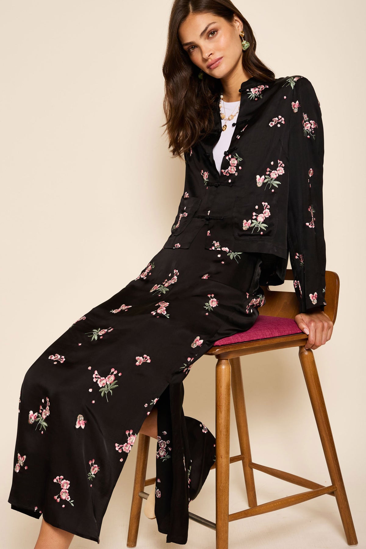 A woman with long brown hair sits on a wooden chair, wearing the Indiya - Blossom Embroidery Pink skirt and matching buttoned jacket. The black outfit has pink floral embroidery, and she gazes at the camera with a relaxed expression.