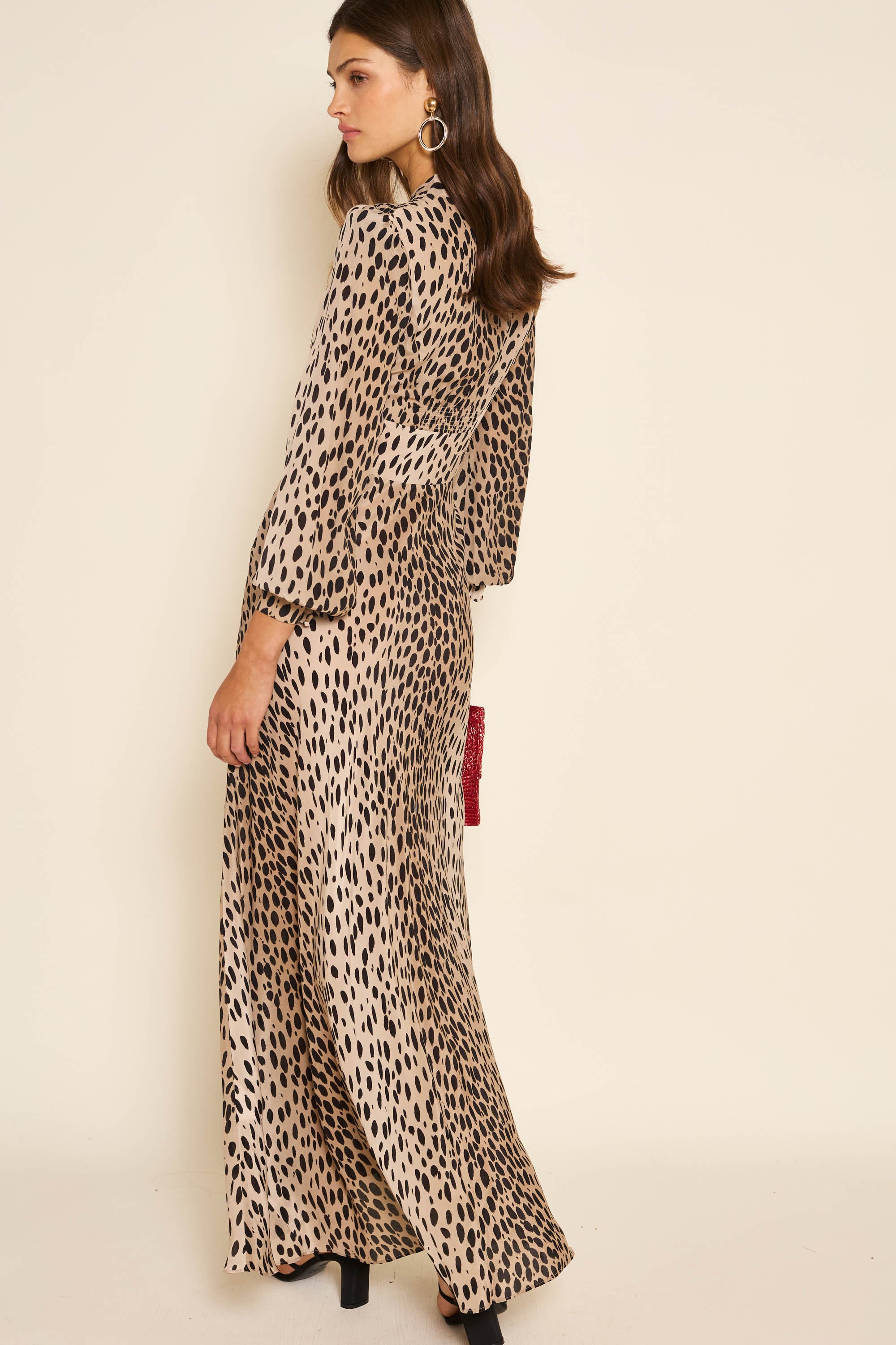 A woman with long brown hair stands sideways against a plain light background, wearing the Emory - Antelope Spot Black dress with black cheetah print, black heels, gold hoop earrings, and holding a small red bag.