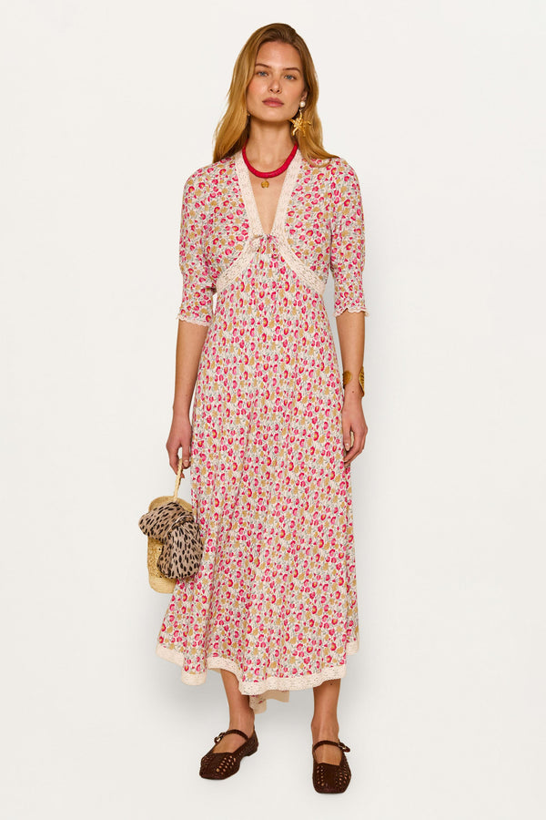 A woman stands against a plain background in the Amina - Shell Pansy Ivory dress, featuring a long floral print and elbow-length sleeves. She accessorizes with a small straw bag, a leopard print detail, and brown woven shoes.