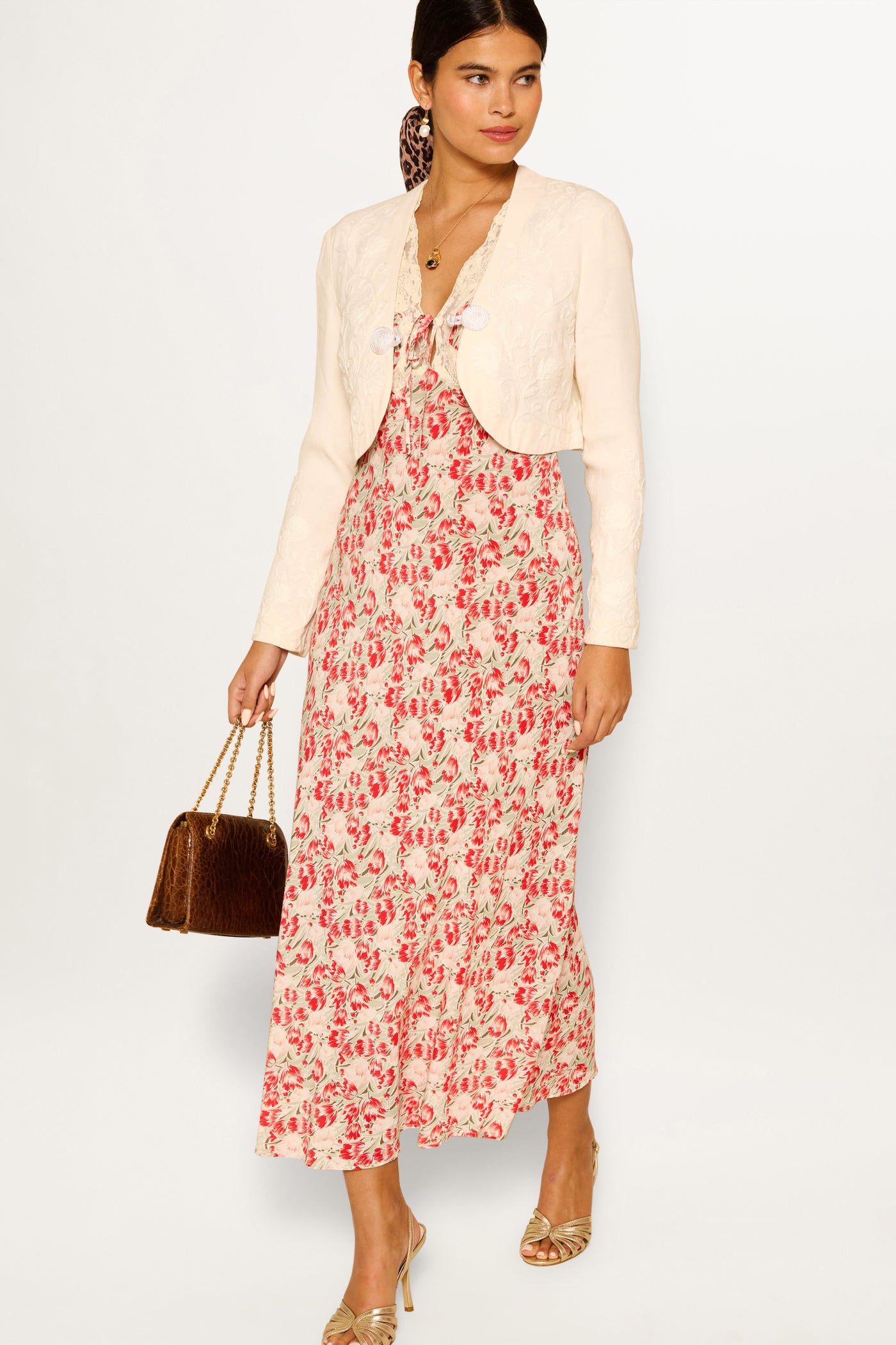 A woman stands against a white background wearing the Amari - Tulip Fields Red dress with a cream cropped jacket, brown handbag, gold open-toe heels, and her hair tied back with a scarf.